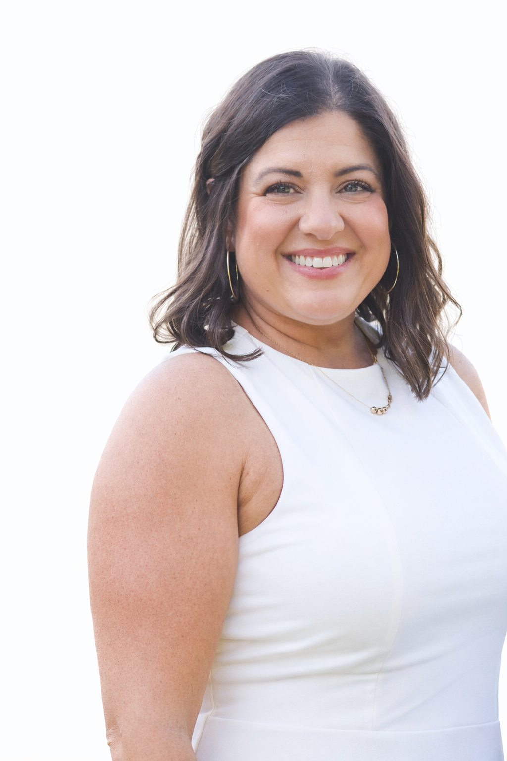 A smiling woman with dark wavy hair, wearing a sleeveless white top, gold hoop earrings, and a gold necklace, standing against a white background.