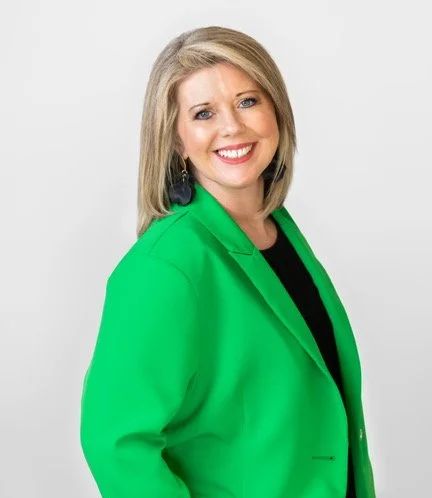 A smiling woman with blonde hair wearing a green blazer and black top, standing against a white background.
