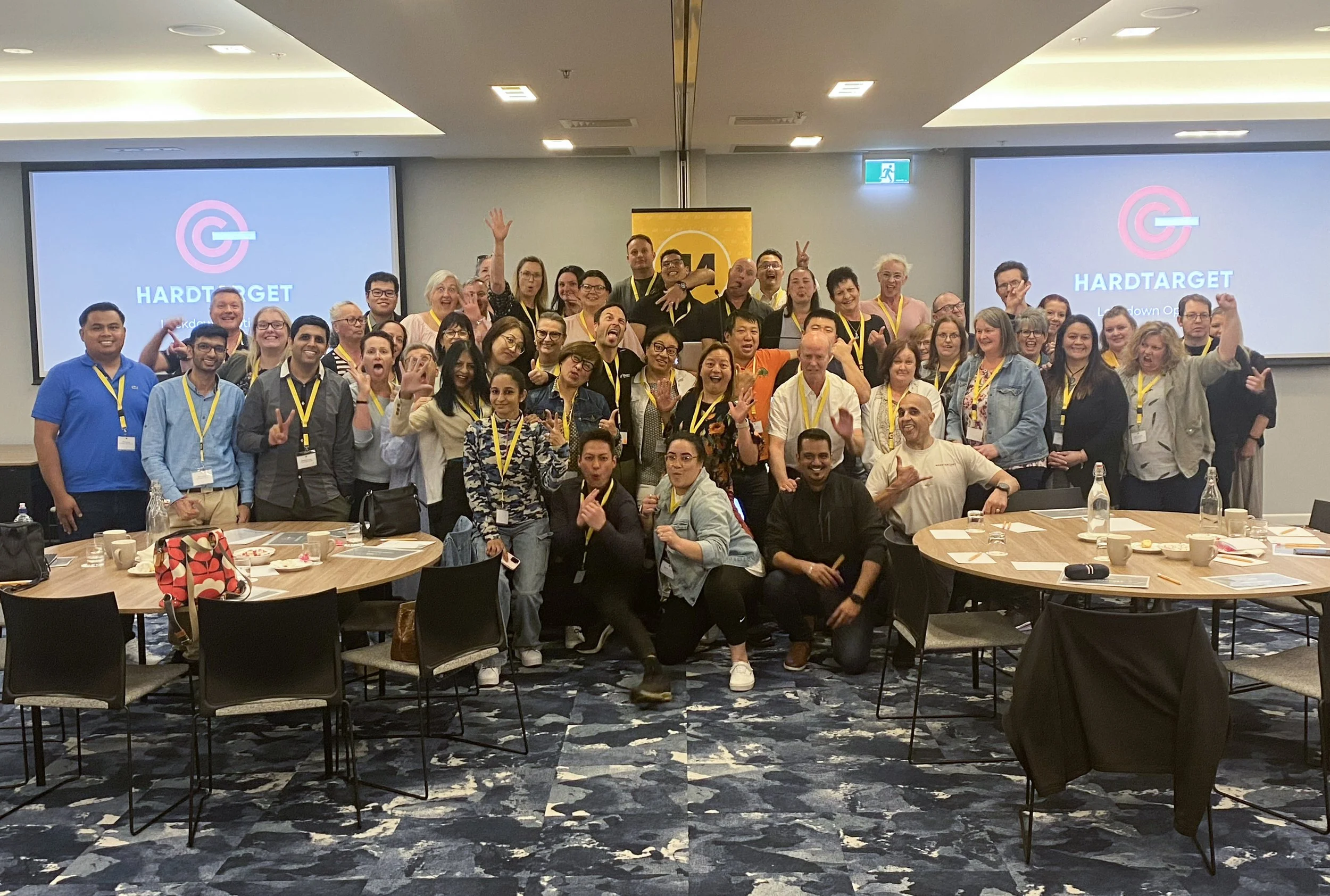 Group of diverse people from AA in a conference room, some standing and some sitting, smiling, with two screens displaying the HARDTARGET logo behind them.