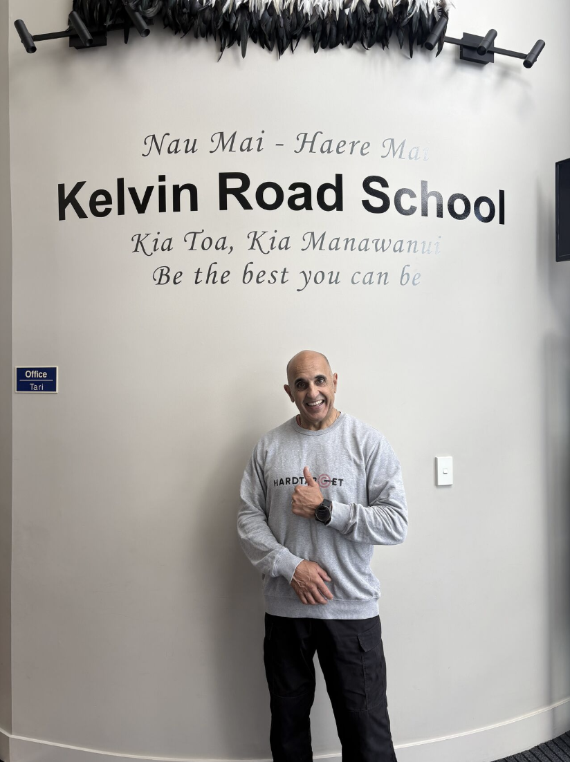 A happy man standing in front of a wall at Kelvin Road School, giving a thumbs-up gesture. The wall has the school's name and motto. There is a sign indicating the office location and a TV monitor.