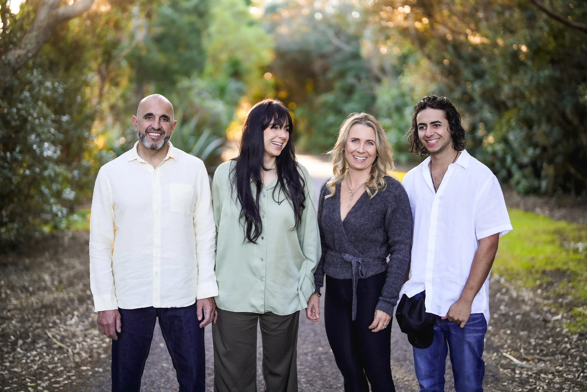 Four people standing outdoors on a dirt path surrounded by trees and greenery, smiling and looking happy.