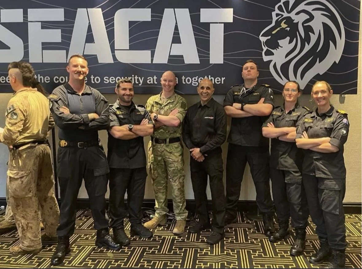 Group of military personnel and security officers standing in front of a banner that reads 'SEACAT,' with a lion's head logo, in a conference or event setting.