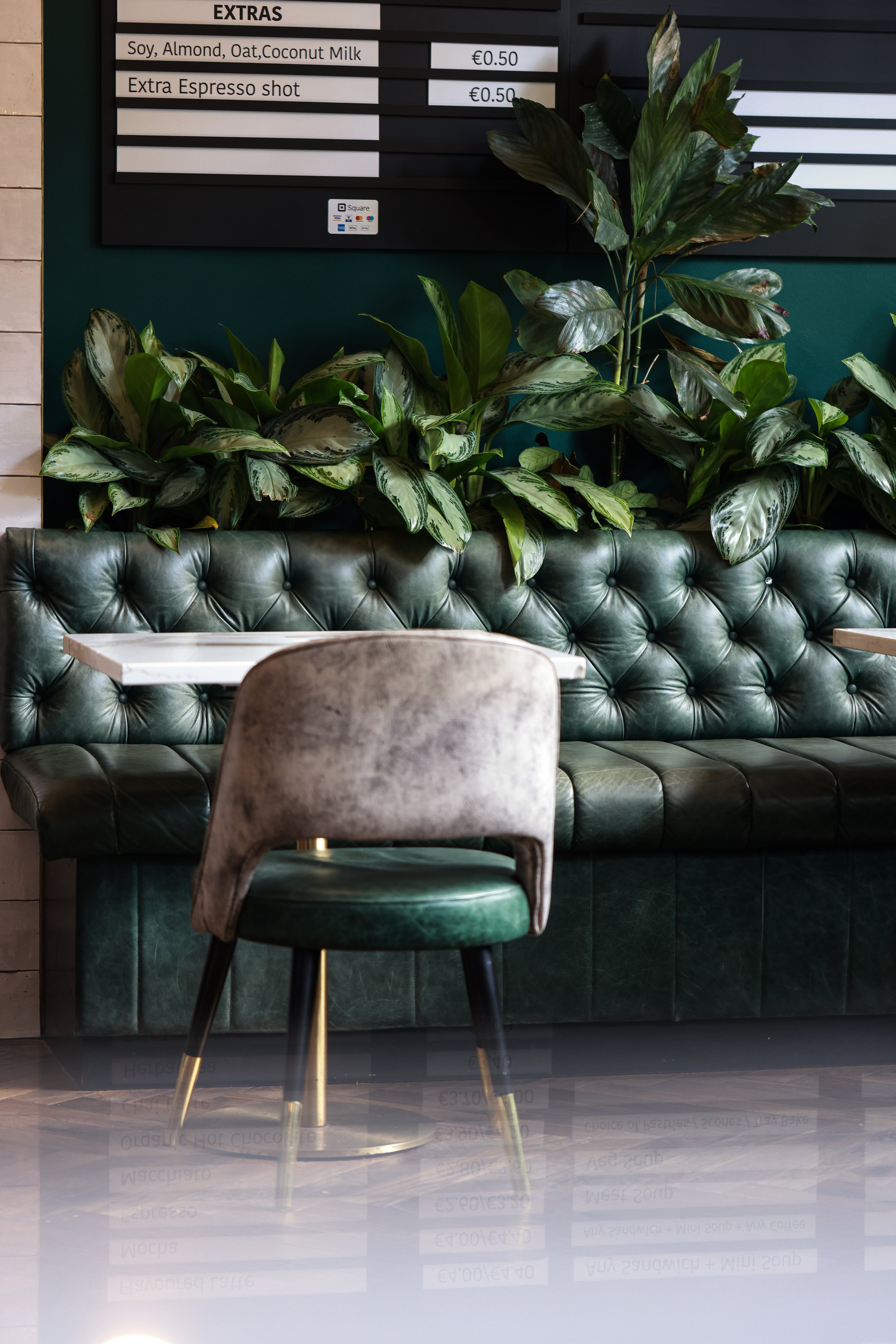 Cozy café interior with green leather seating, a chair, decorative plants, and a menu board displaying drink extras.