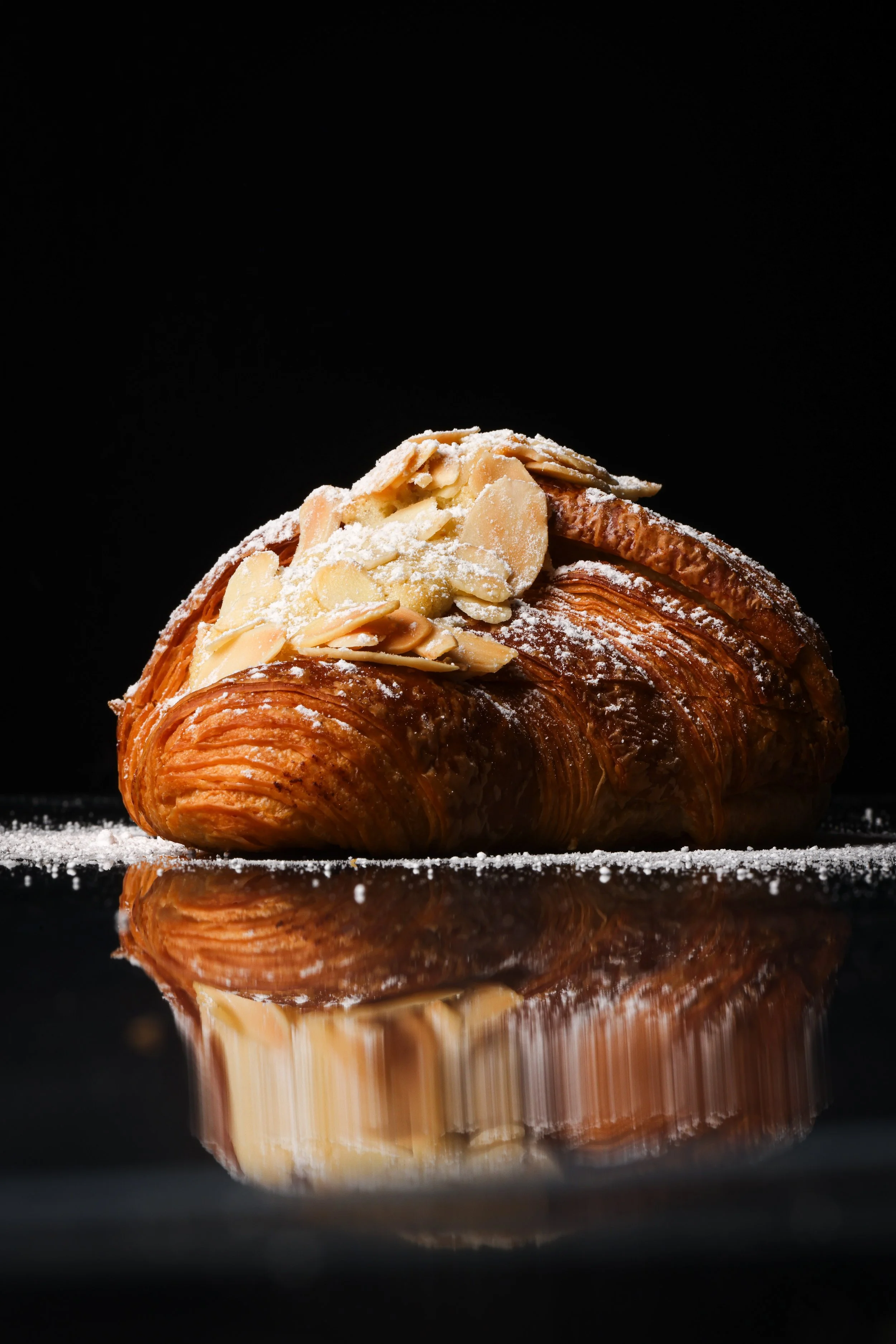 Close-up of an almond croissant with powdered sugar on a reflective surface against a black background.