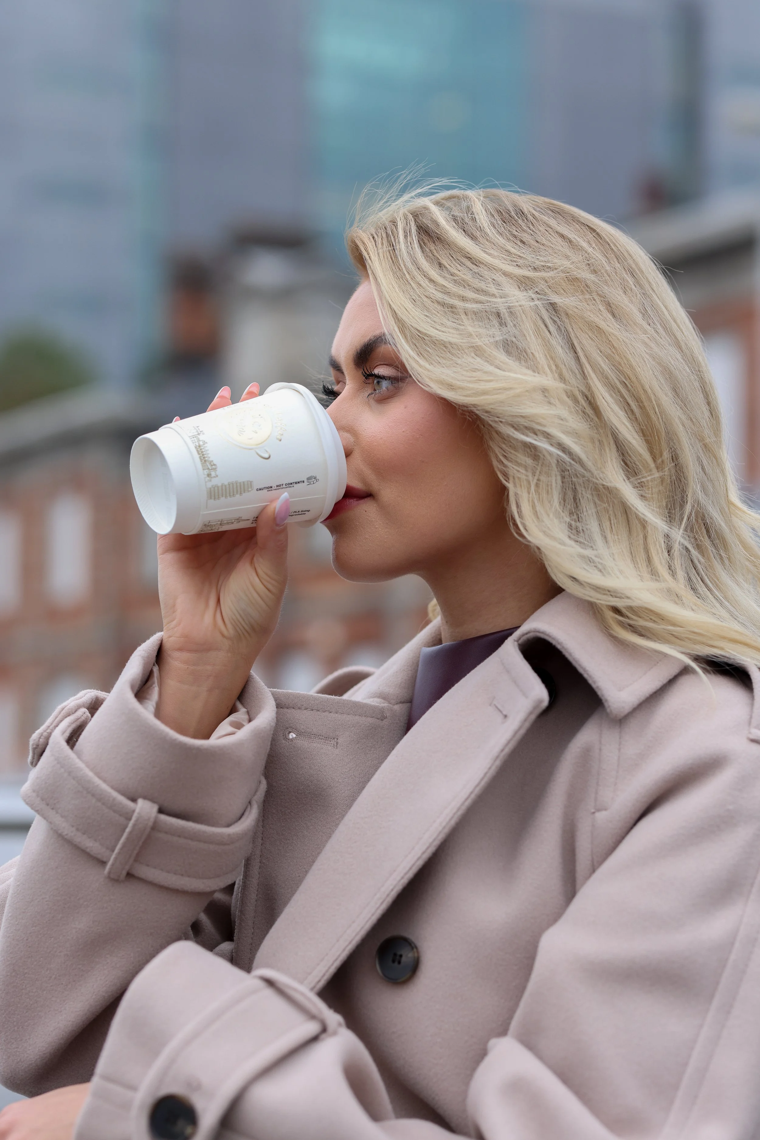 Woman drinking from a paper cup outdoors, wearing a beige coat.