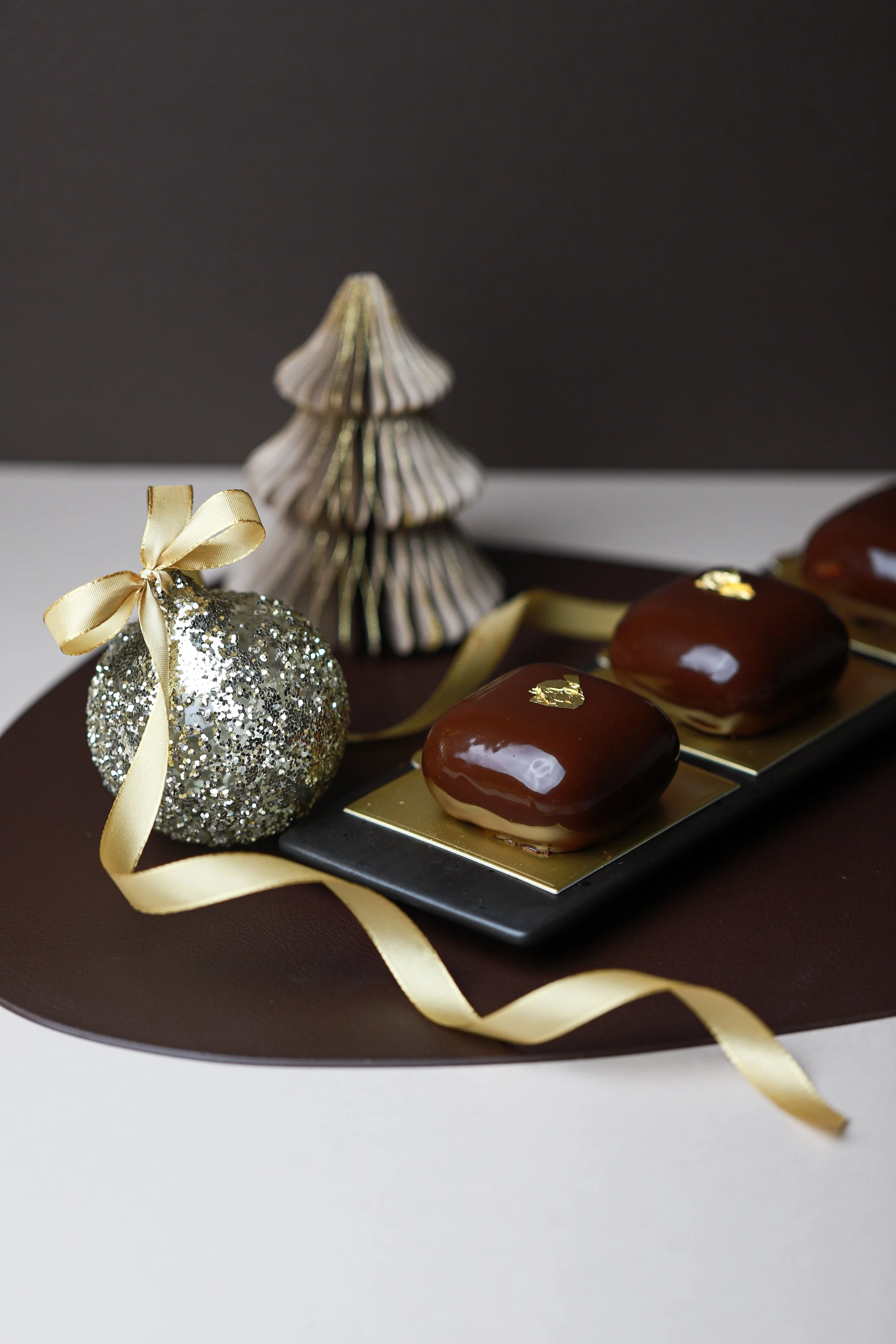 Three chocolate glazed pastries with gold leaf decoration on a black tray, surrounded by a glittery ornament with a gold ribbon and a decorative paper Christmas tree.