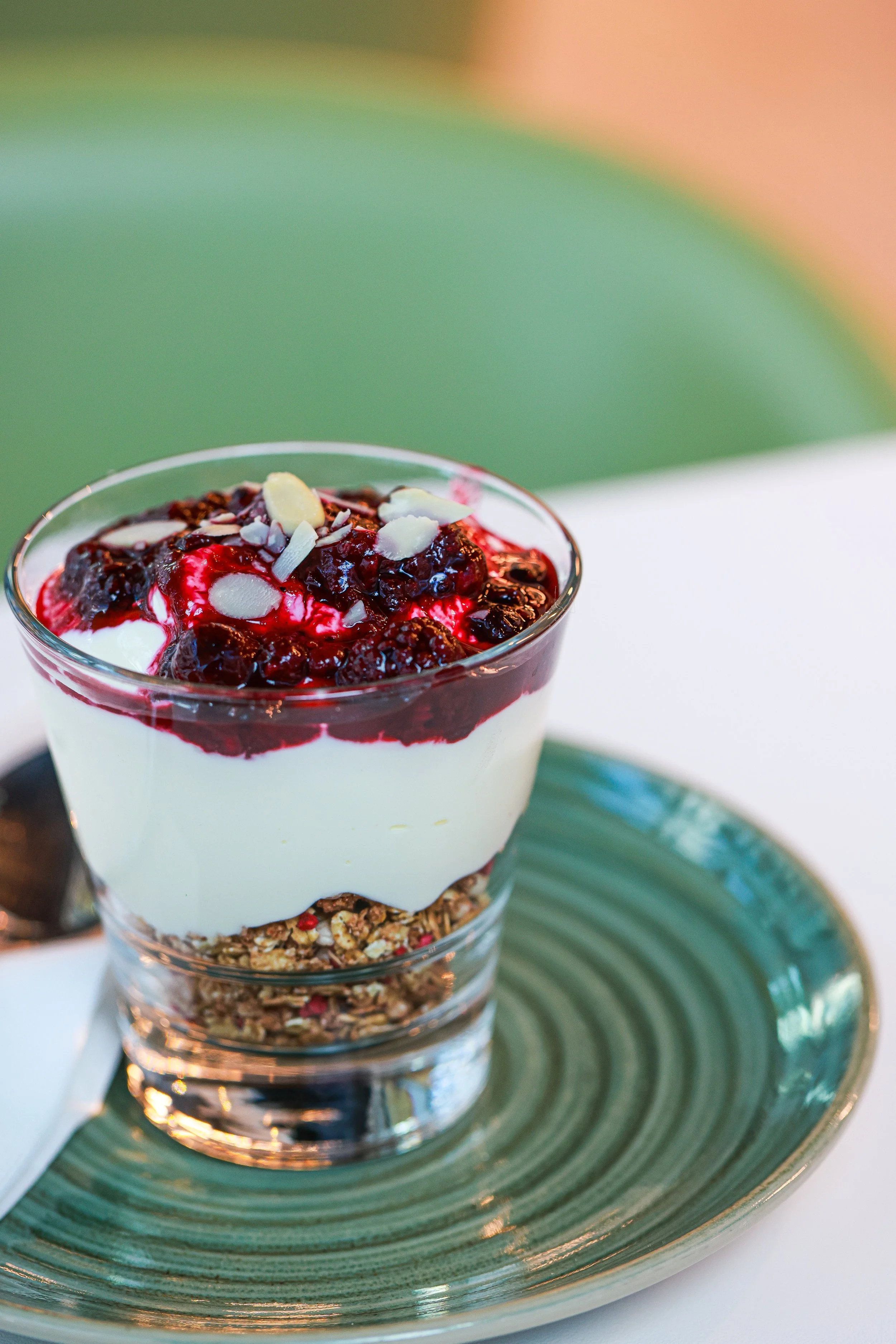 Glass of layered yogurt parfait with granola at the bottom, creamy yogurt in the middle, and berry compote topped with almond slices on top, served on a green plate.
