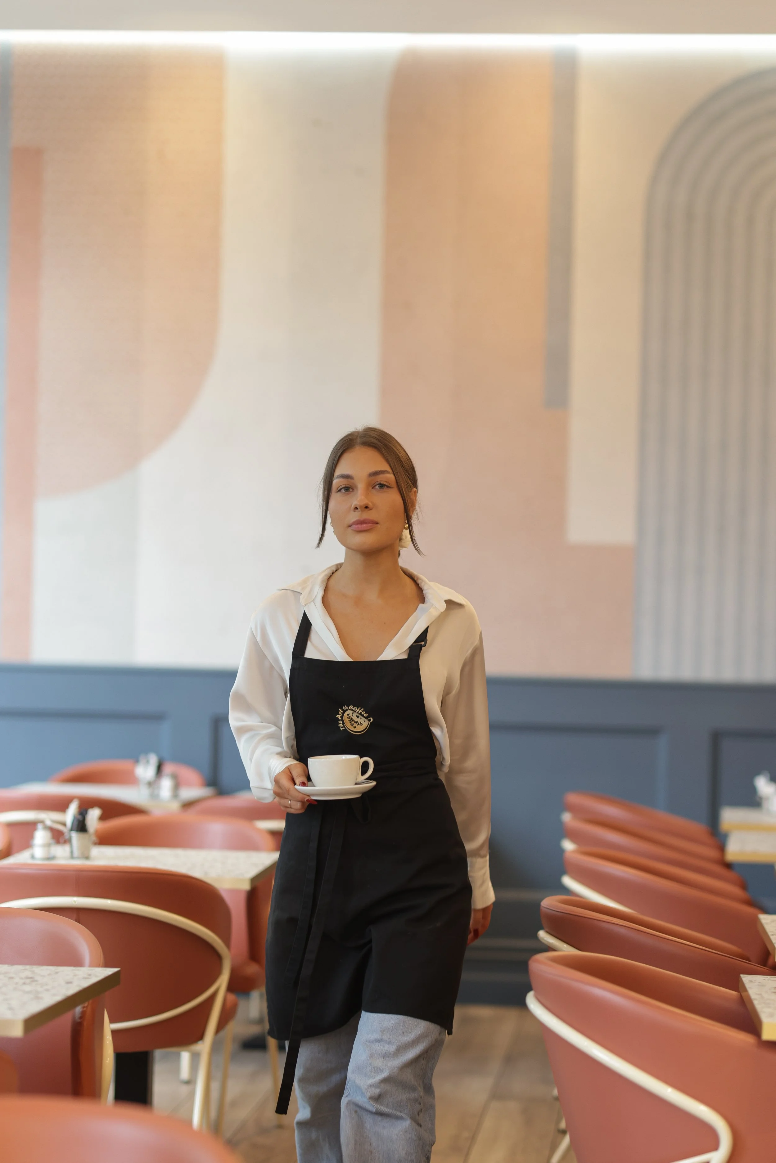 Waitress in a modern cafe holding a cup of coffee, wearing a white shirt and black apron, with salmon-colored chairs and abstract wall art in the background.