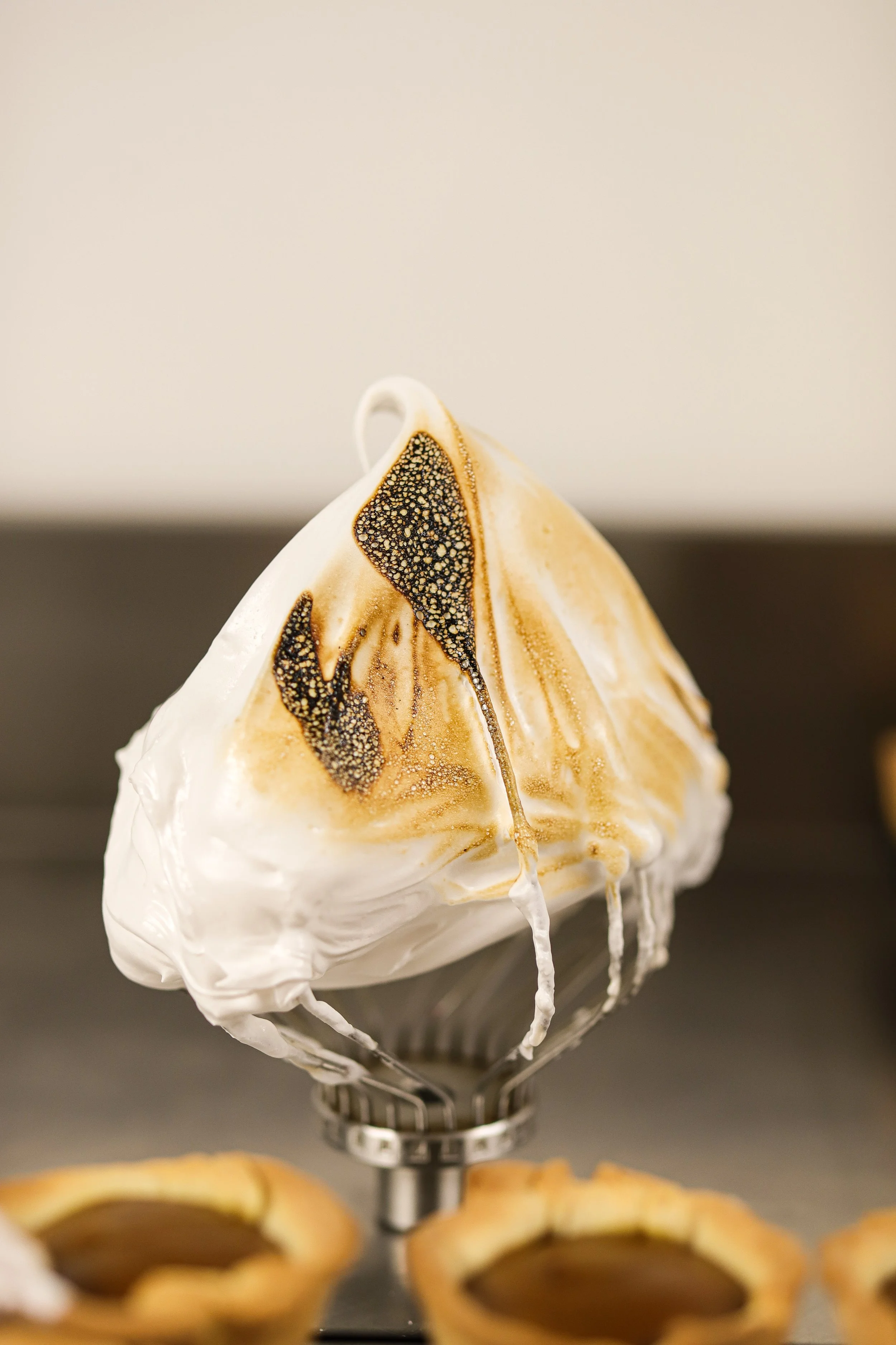 Whipped cream with toasted peaks on a mixer whisk, with pastries in the background.