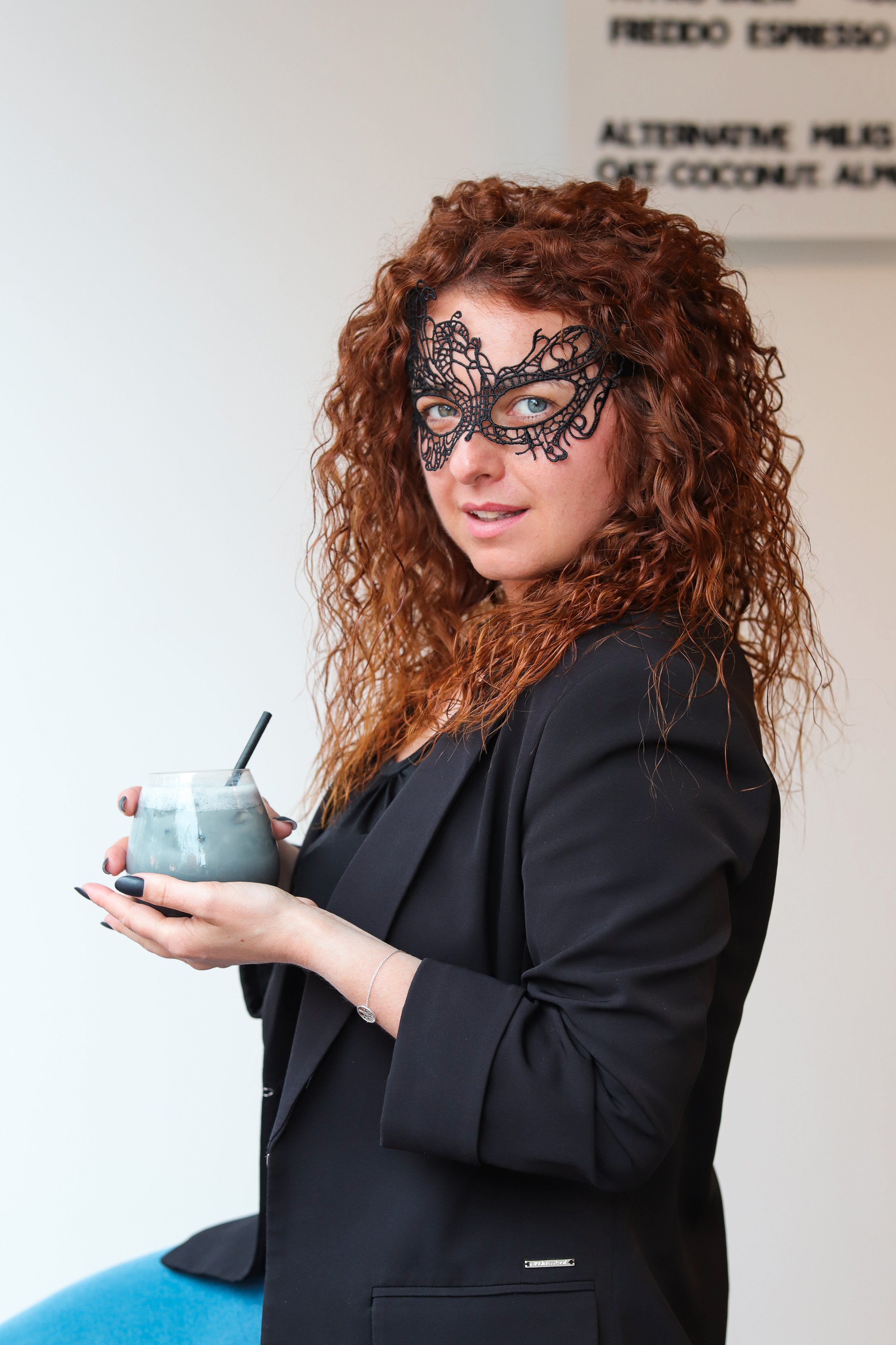 A woman with curly red hair wearing a black lace mask and holding a glass of blue beverage with a straw, dressed in a black blazer.