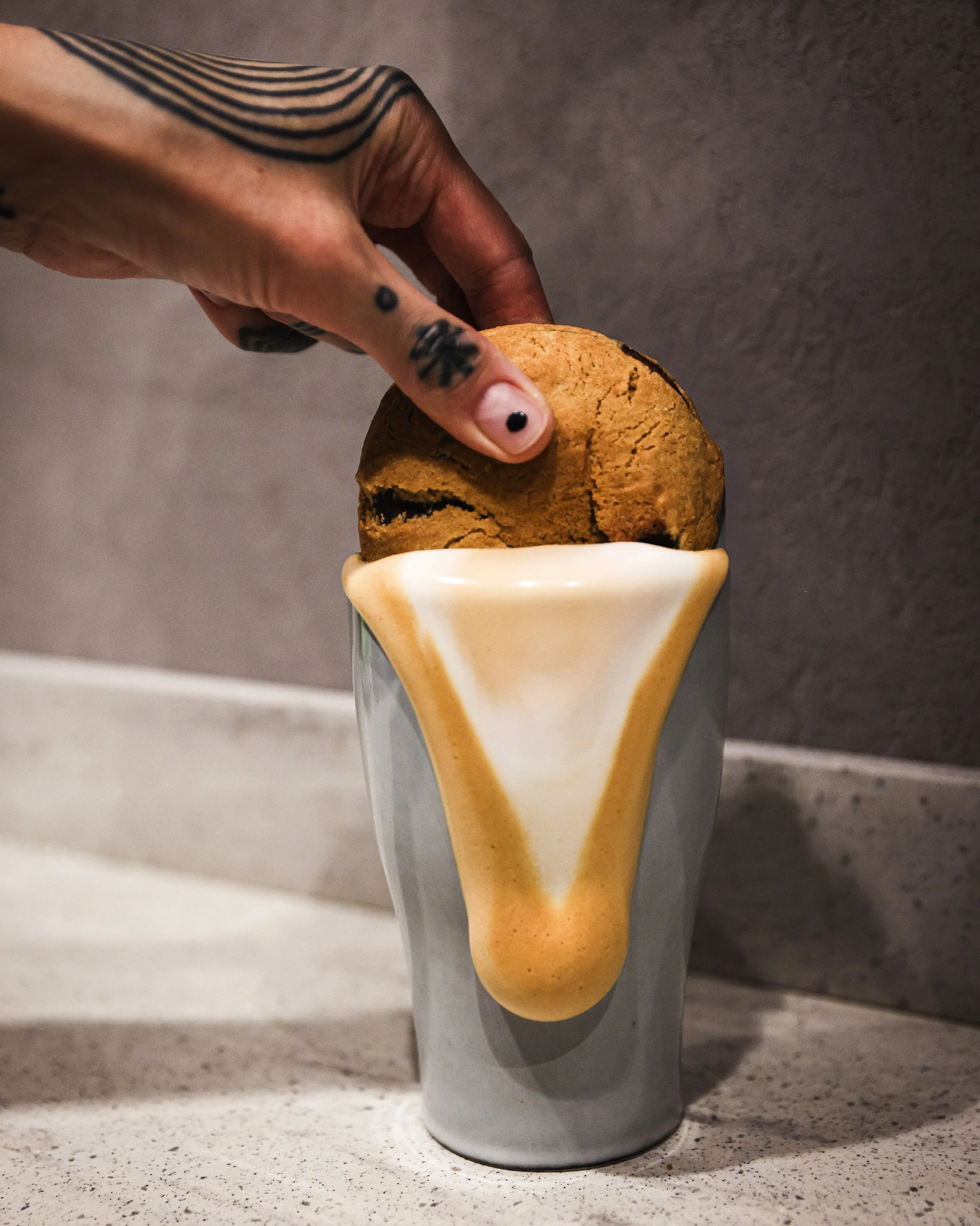 A hand with tattoos holding a chocolate chip cookie above a ceramic mug with a unique drip design, suggesting a coffee theme.