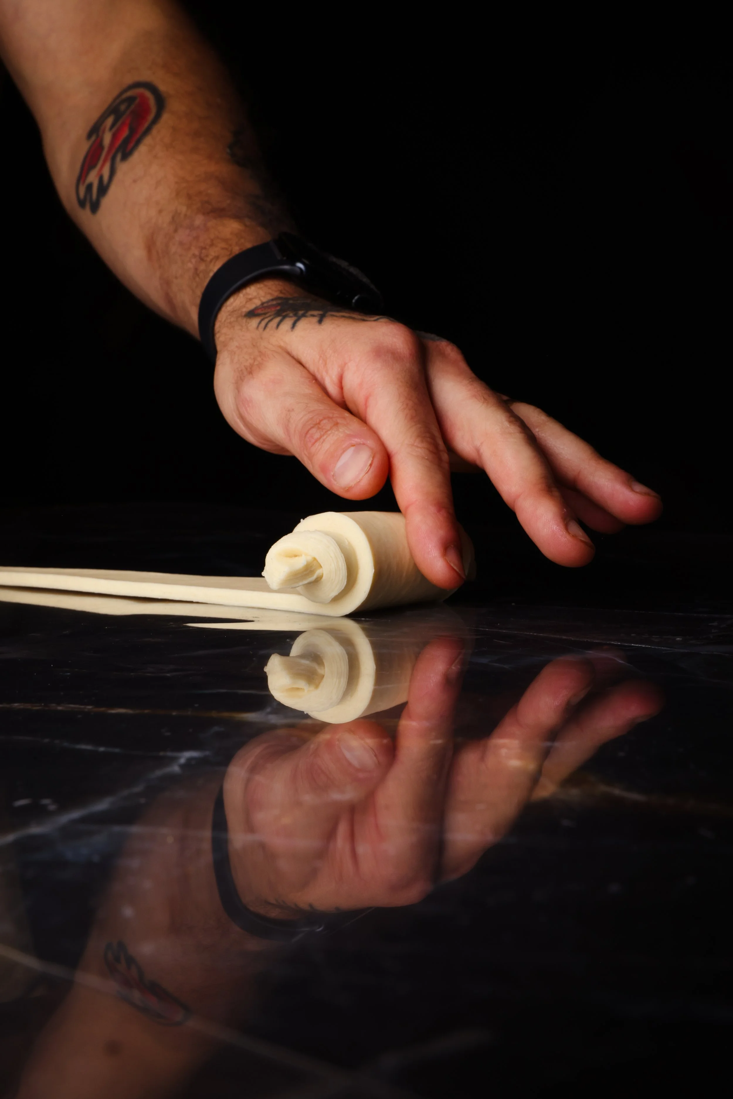 Hand rolling thin strips of dough on a black surface, with a tattoo visible on the arm.