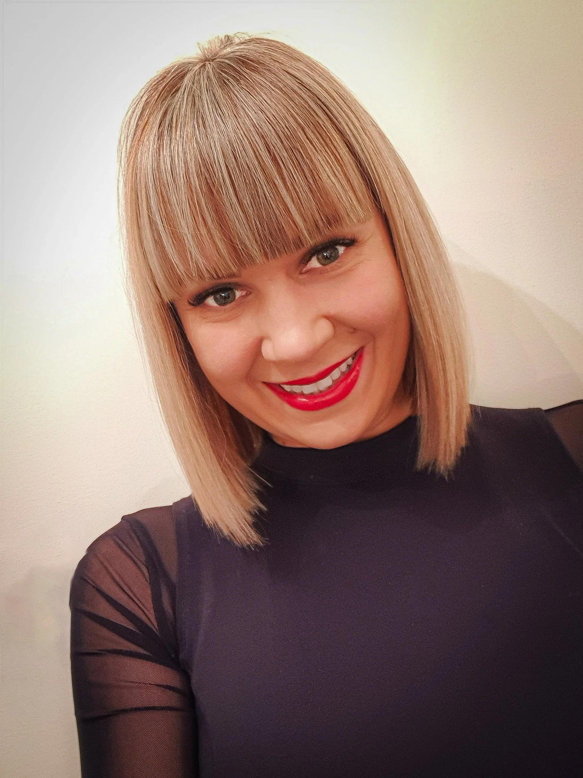 Smiling person with straight blonde hair and bangs, wearing a black top.