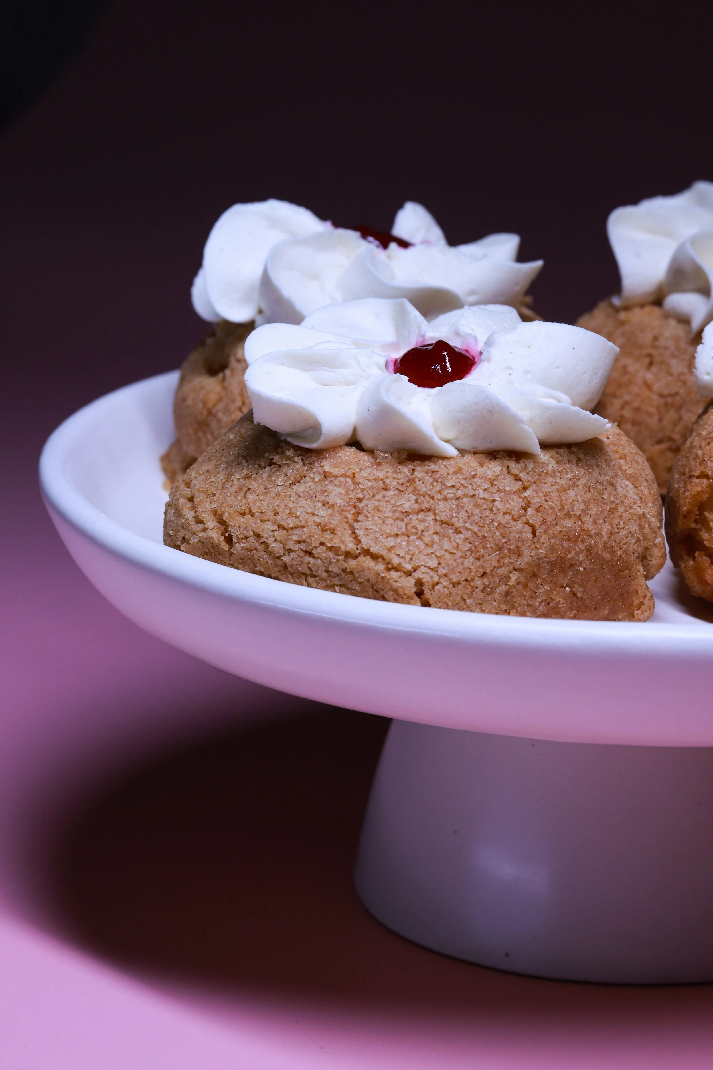Cookies topped with whipped cream and red jam on a white cake stand, set against a pink background.