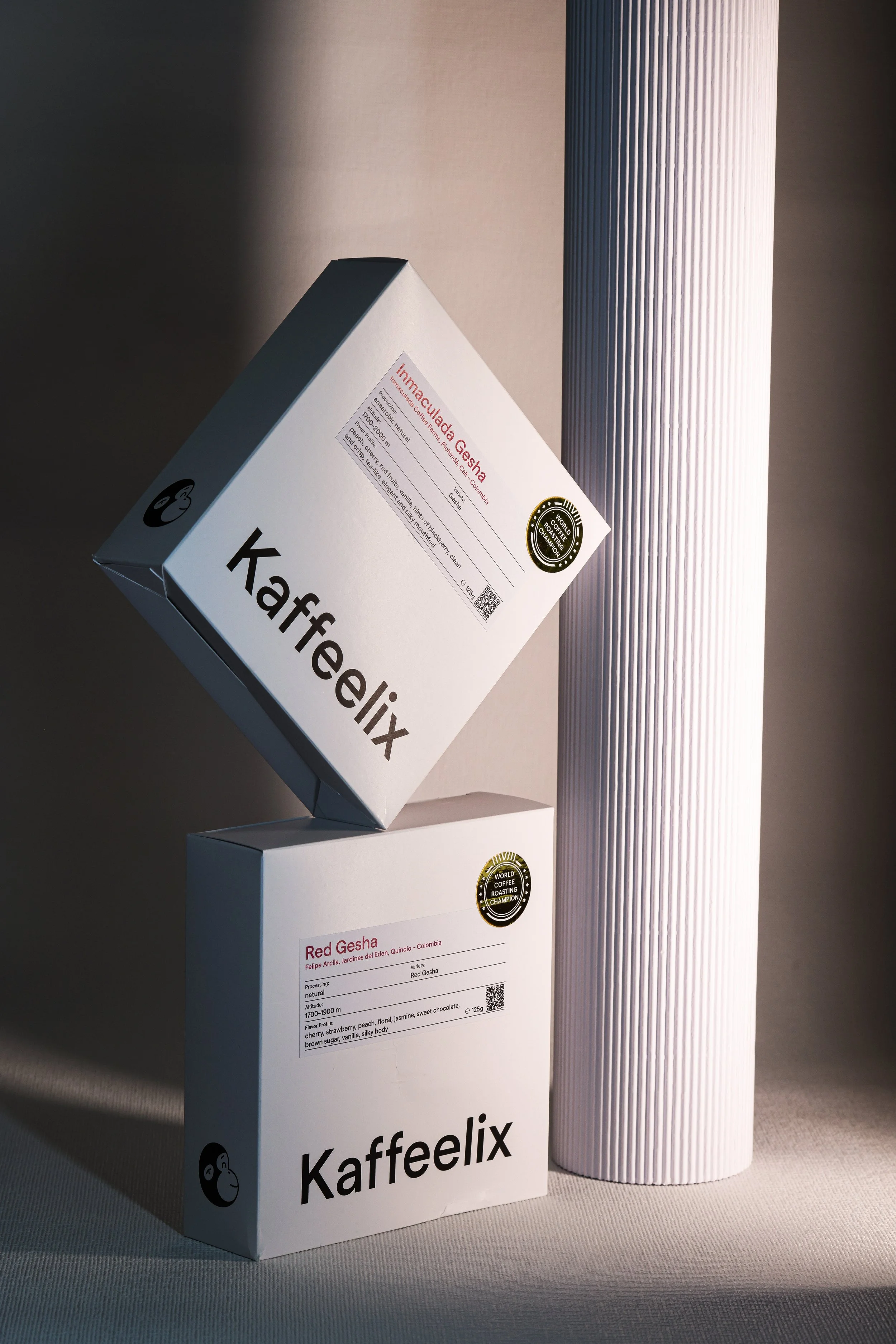Two white coffee packaging boxes labeled 'Kaffeelix' with different types of Gesha coffee, alongside a vertical textured white cylinder.