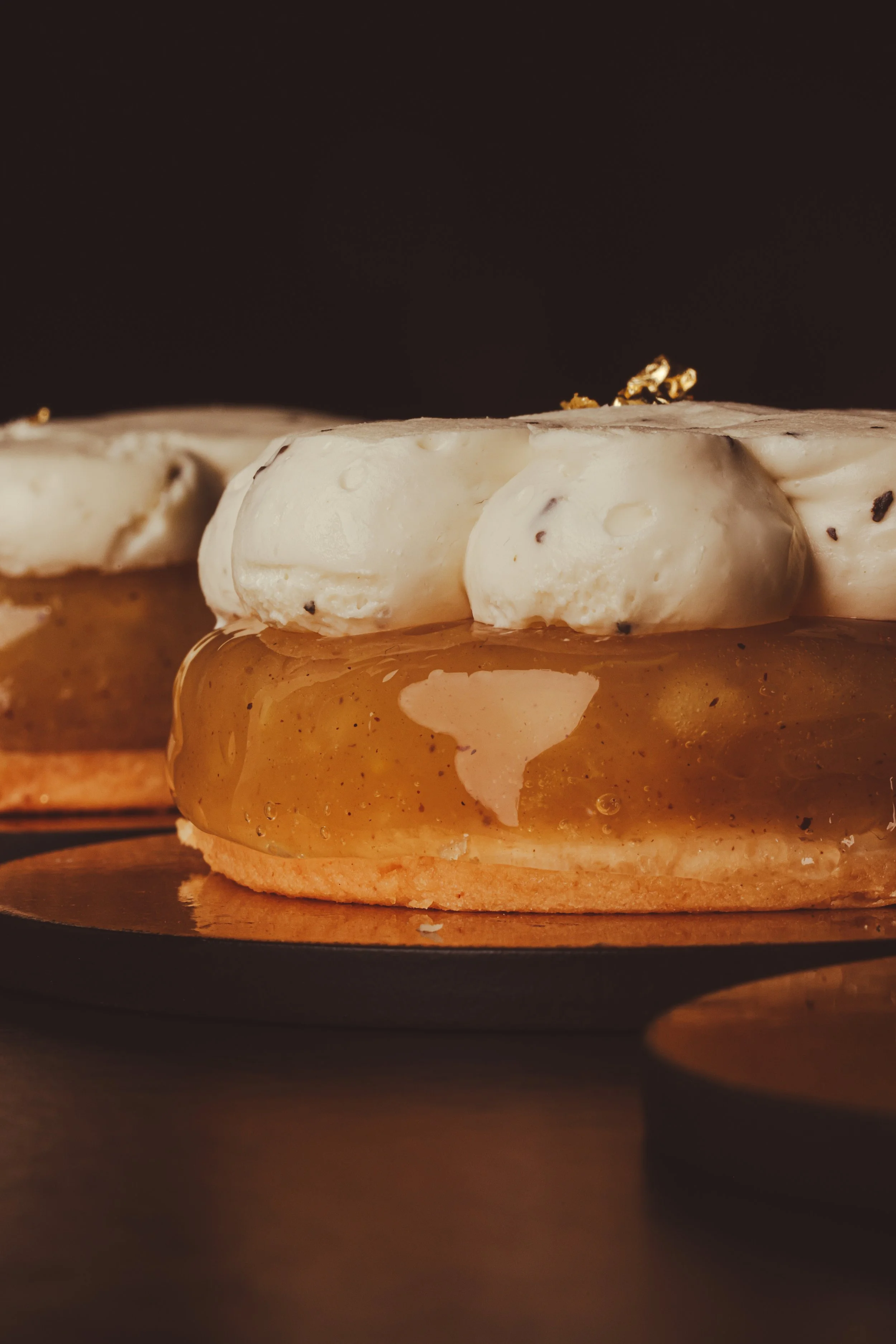 Close-up of a gourmet dessert with a gel-like layer and whipped topping, possibly adorned with gold leaf, set on a round biscuit base.