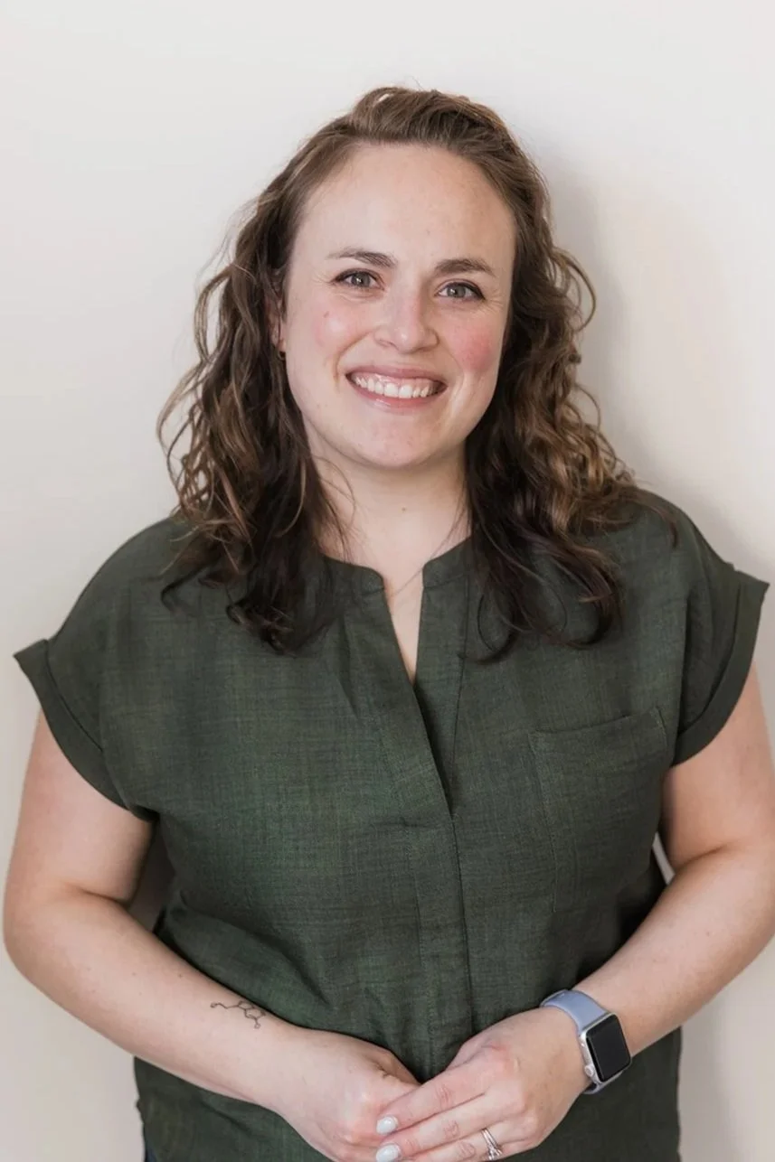 A smiling woman with shoulder-length brown hair, wearing a black top and beige cardigan, standing in front of a white brick wall.