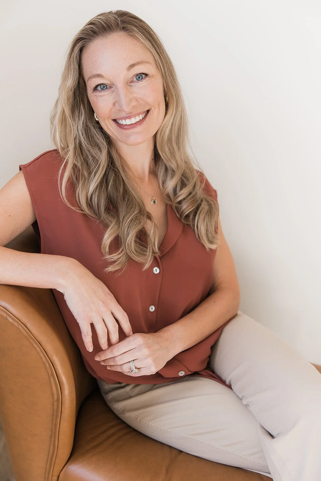 Portrait of a smiling woman with long blonde hair, wearing a light gray blazer and a gray top, standing in front of a white brick wall.
