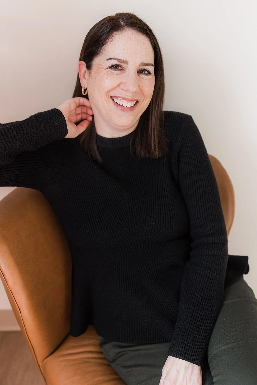 A woman with dark hair, wearing a denim jacket and gold hoop earrings, smiling in front of a plain light-colored background.