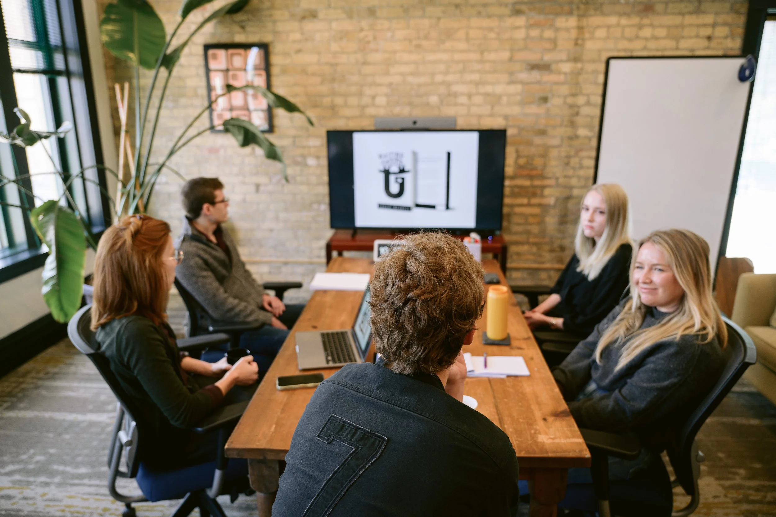 People seated around a meeting table in a conference room with a large screen displaying a logo and a whiteboard.