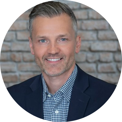 A professional man with short gray hair, blue eyes, and a beard, wearing a navy blazer and checkered shirt, standing in front of a brick wall.