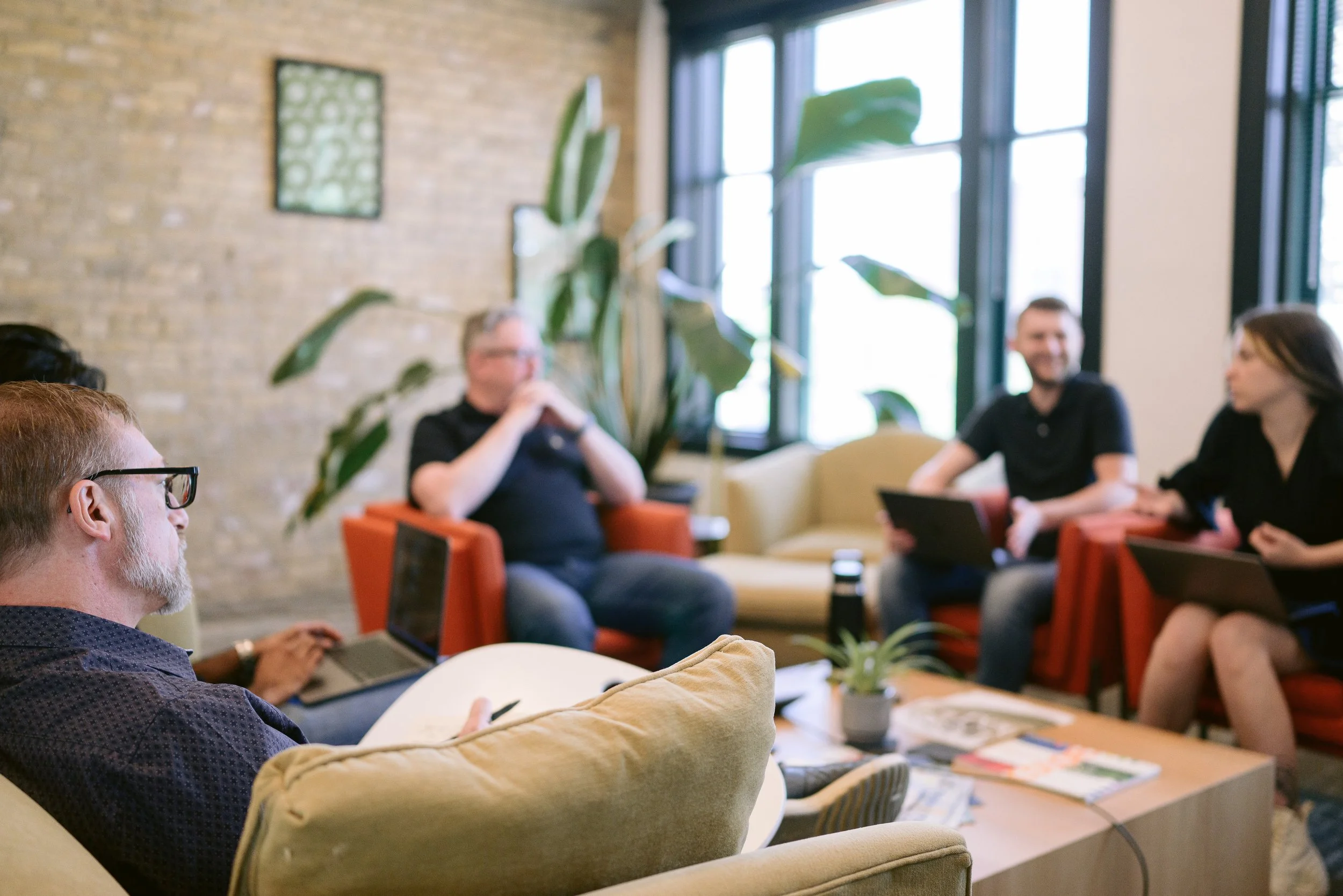 A group of people in a casual meeting room with large windows, sitting on colorful couches, using laptops and tablets, with a large plant and abstract wall art in the background.