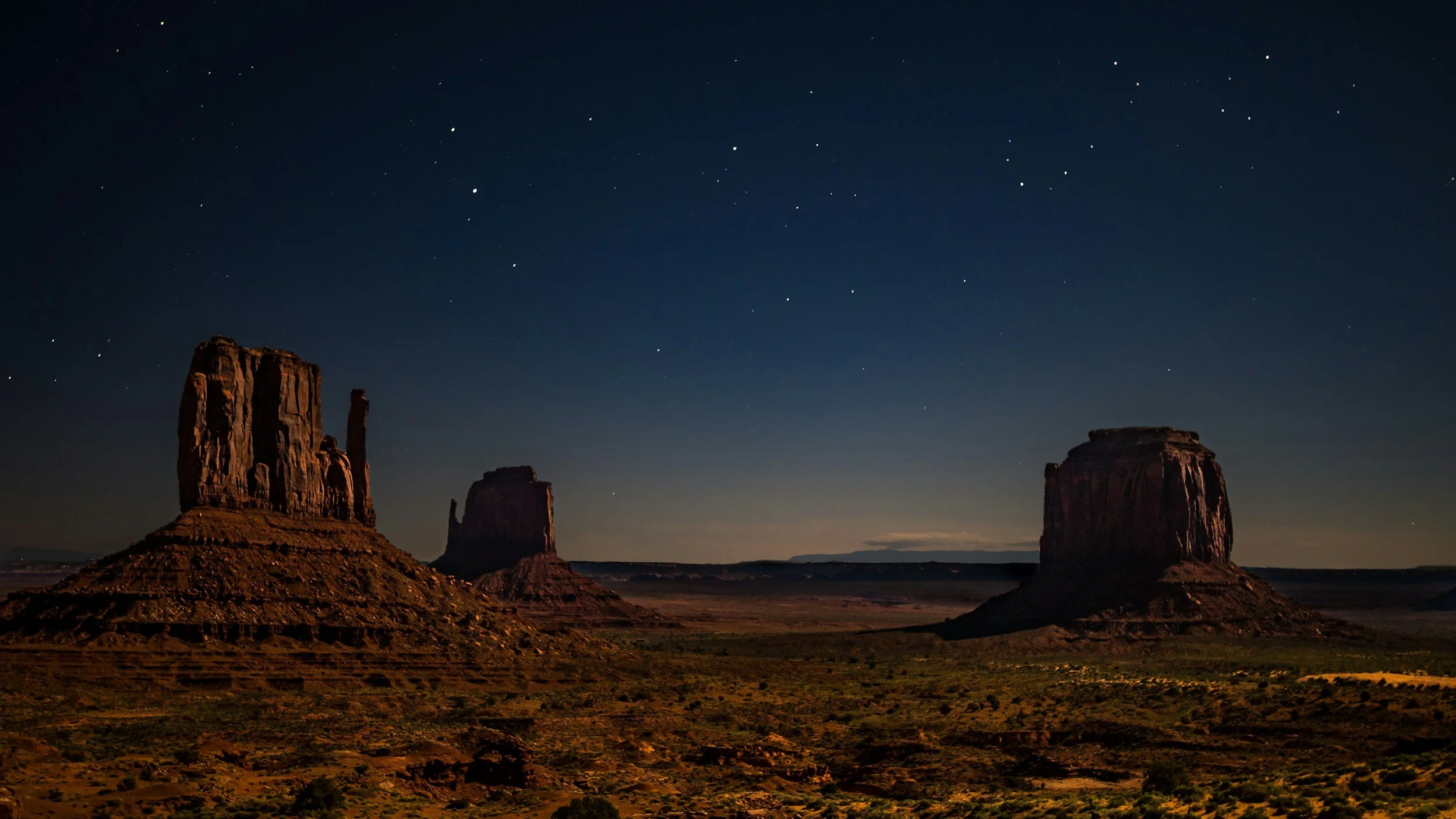 Nighttime view of Monument Valley with three large buttes and stars shining in the sky.