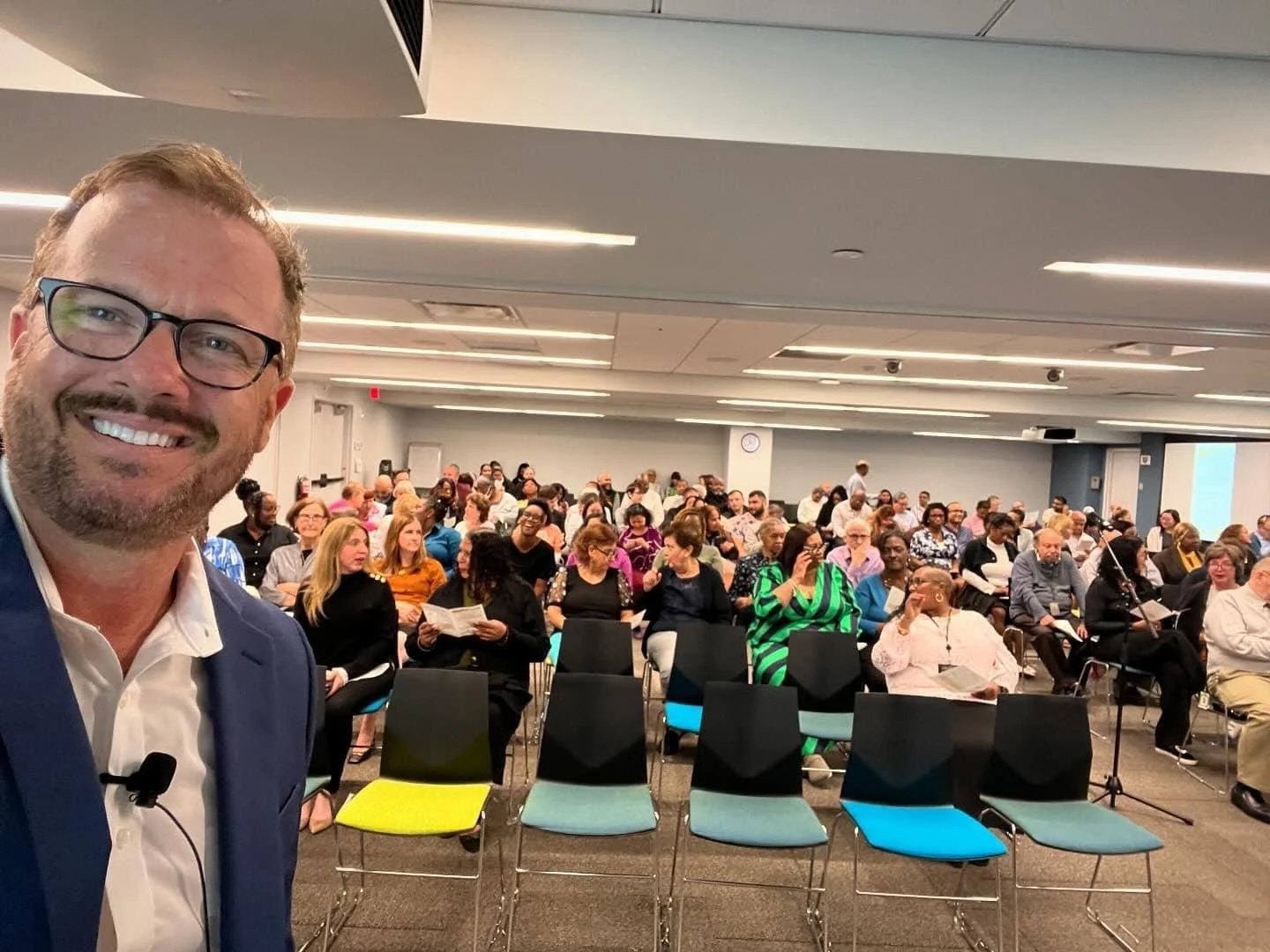 A smiling man in a suit and glasses taking a selfie in front of an audience in a conference room.