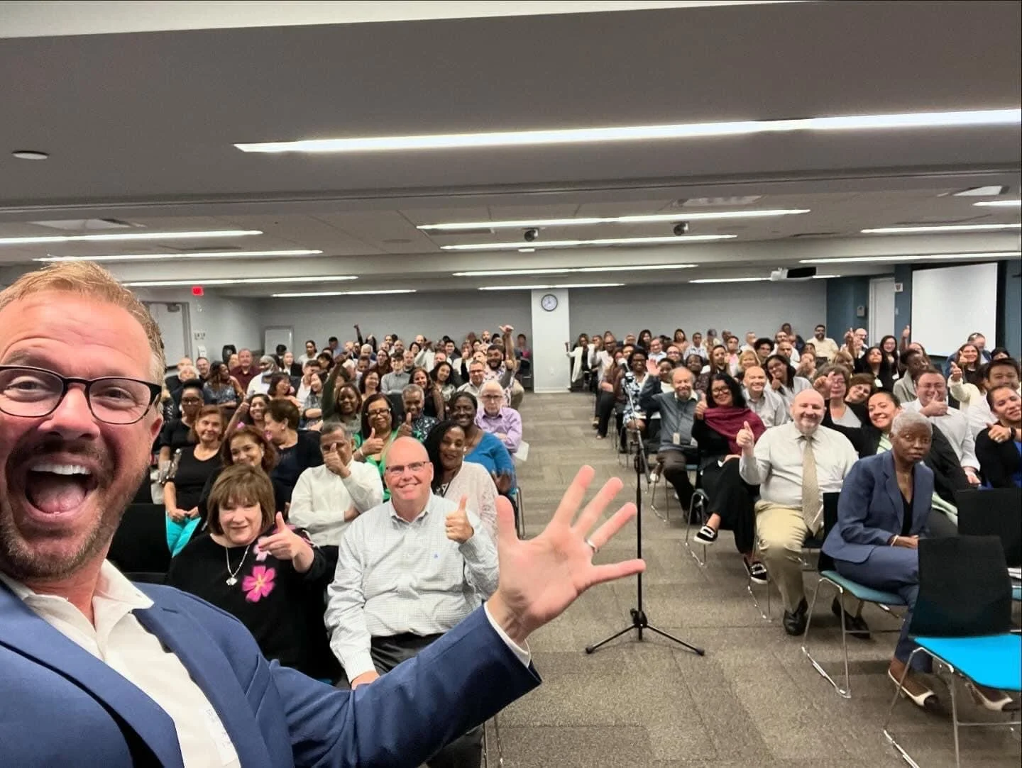 A man in a blue suit and glasses taking a selfie in front of a large diverse audience in a conference room, many of whom are smiling and giving thumbs up.