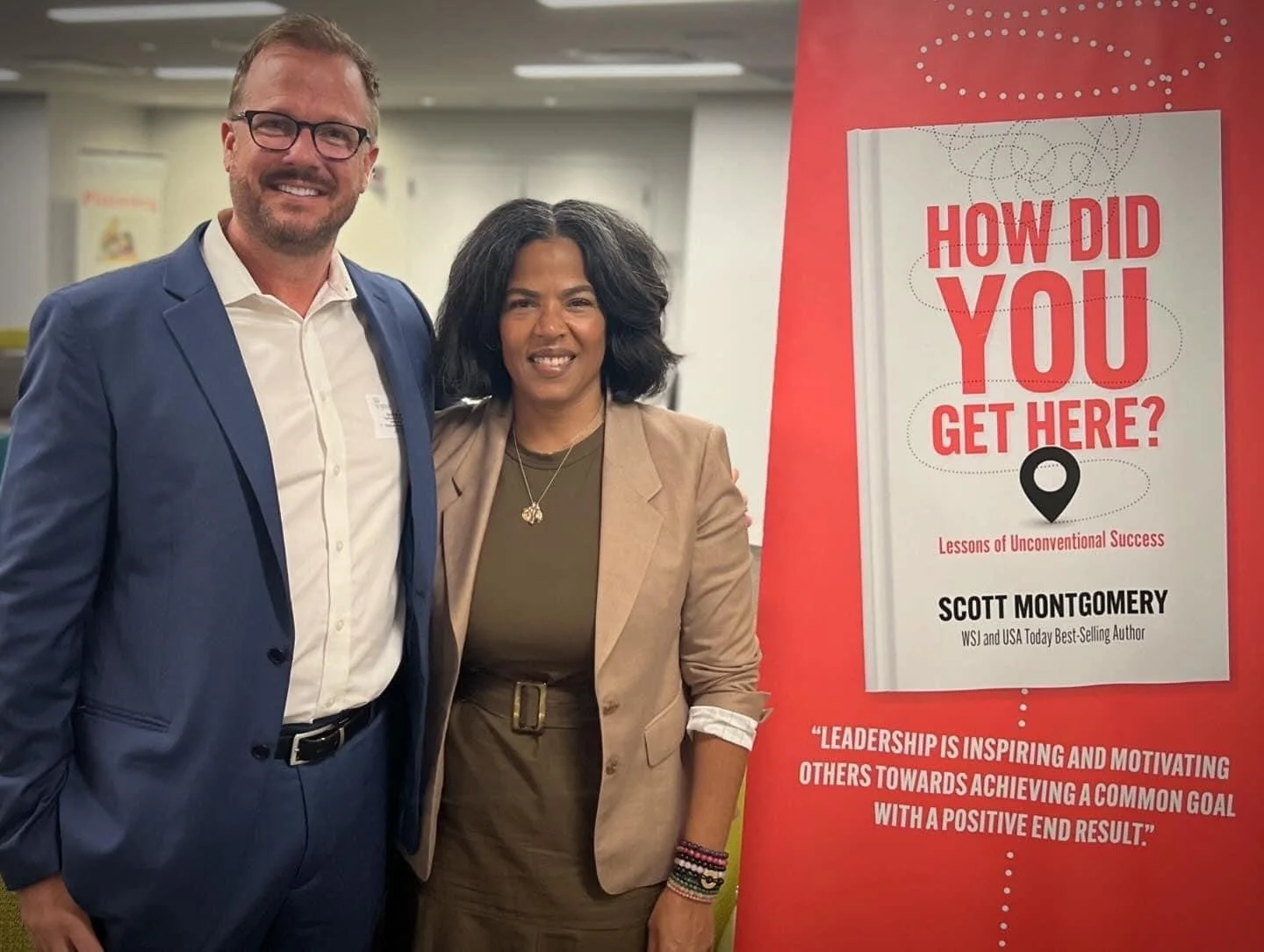 A man and a woman standing together at a book event, smiling. There is a book cover titled 'How Did You Get Here?' by Scott Montgomery displayed beside them.