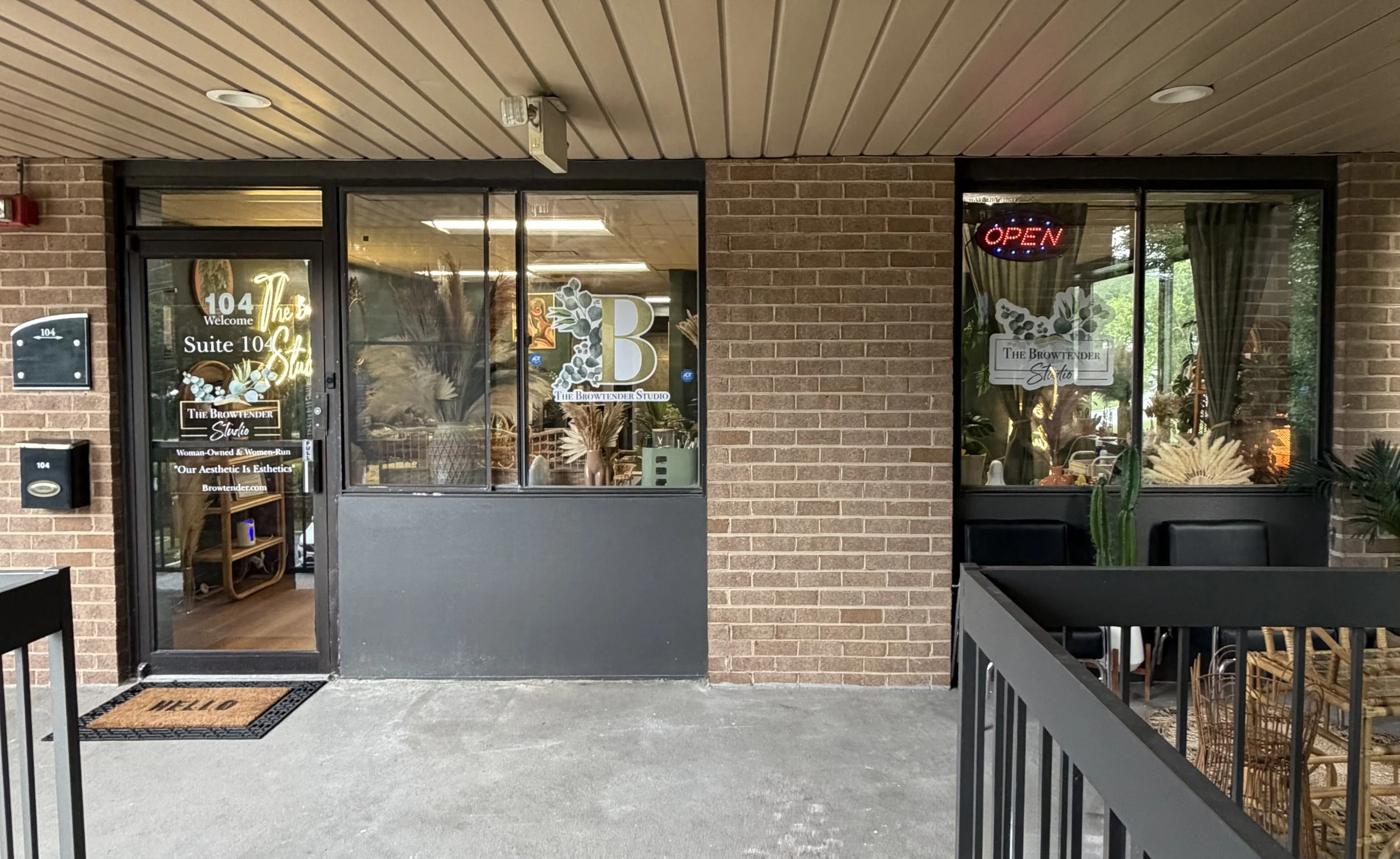 The storefront of The Browtender Studio at 104, with glass doors and windows showing interior decor and plants, an 'OPEN' sign, and a brick exterior wall.