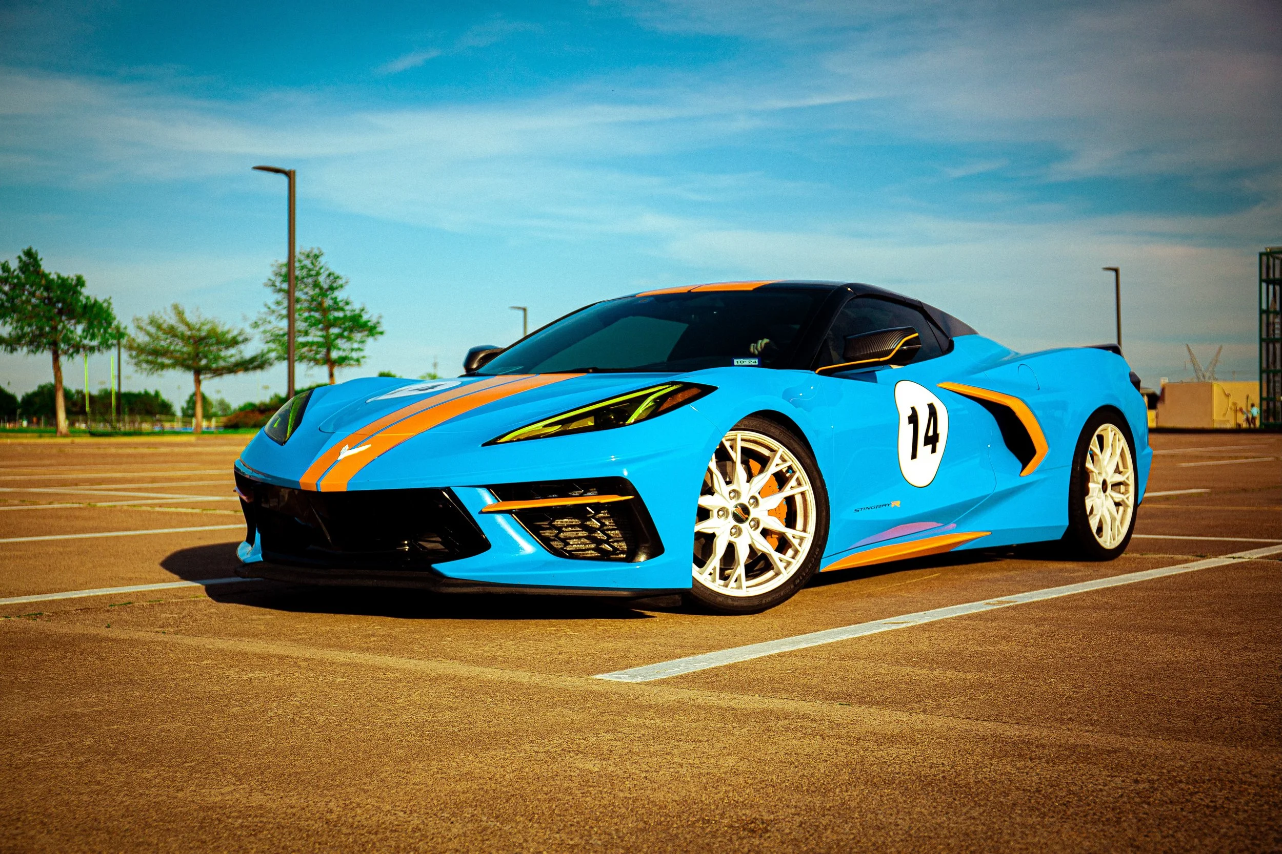 A blue sports car with racing stripes and the number 14 on the side, parked in an empty parking lot on a clear day.