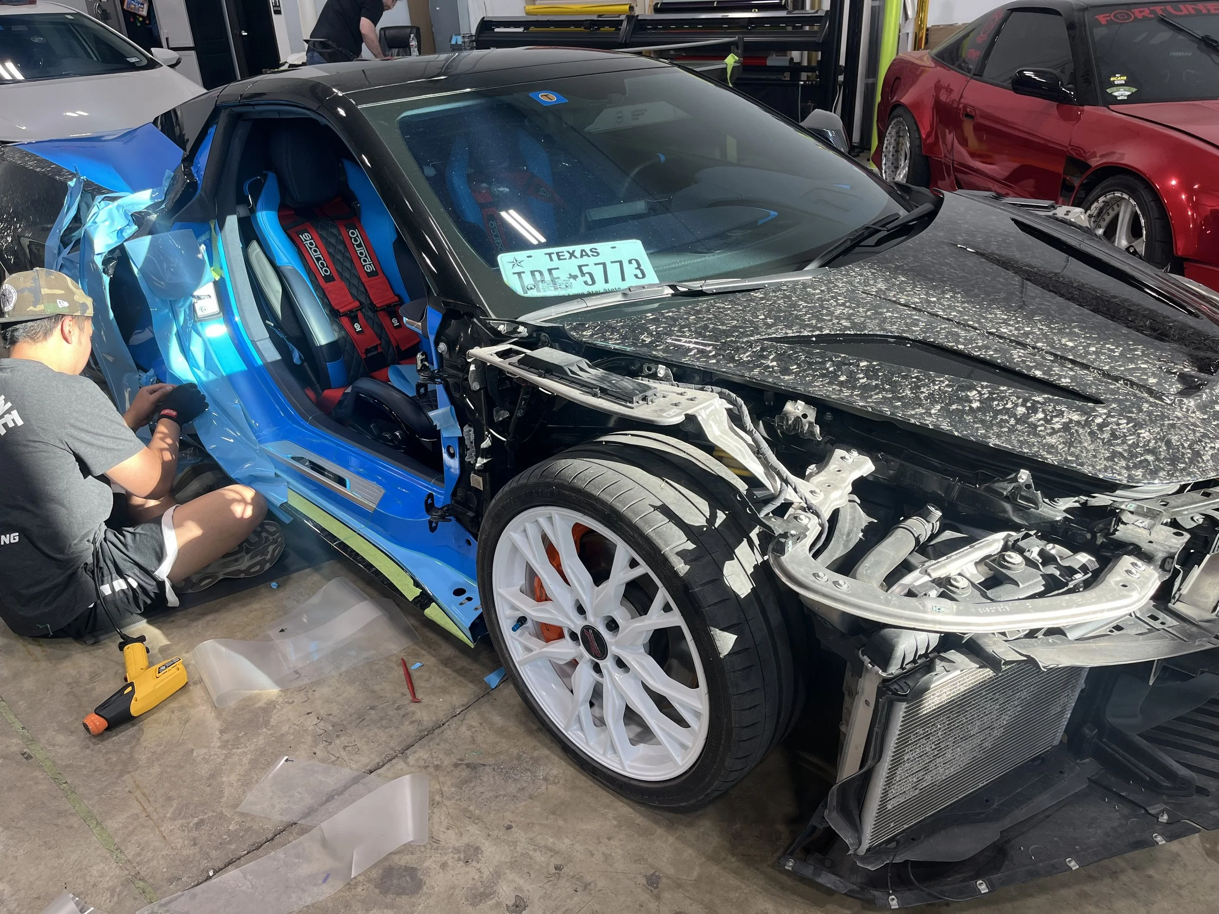 A partially disassembled black sports car with a damaged front end, located in a garage or workshop setting. Inside, racing seats and harnesses are visible, and a person is working on the vehicle. Multiple cars and tools are present in the background.