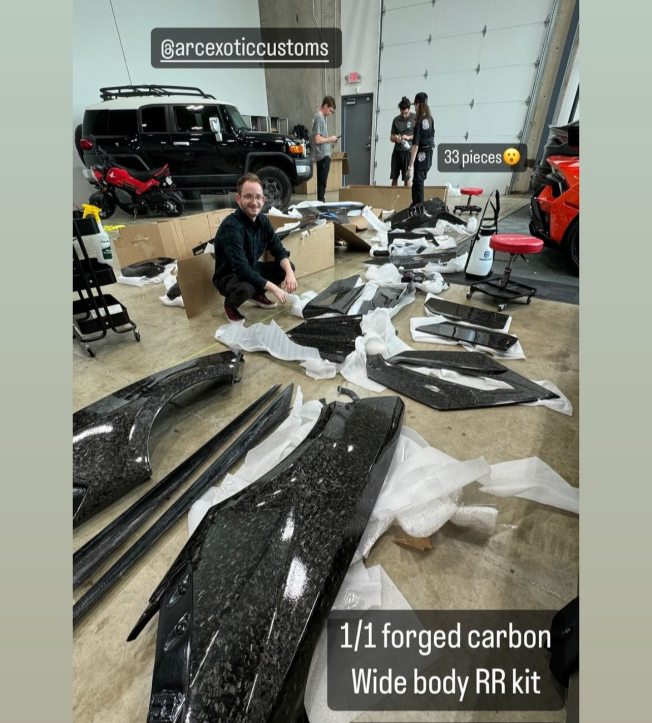 People working in an industrial space with car parts, including a black asphalt-colored carbon fiber wide body kit for a car, some unassembled parts, and various tools. One person is kneeling in the foreground, smiling, next to the assembly. In the background, others are working or talking, with a black vehicle and a red sports car visible.