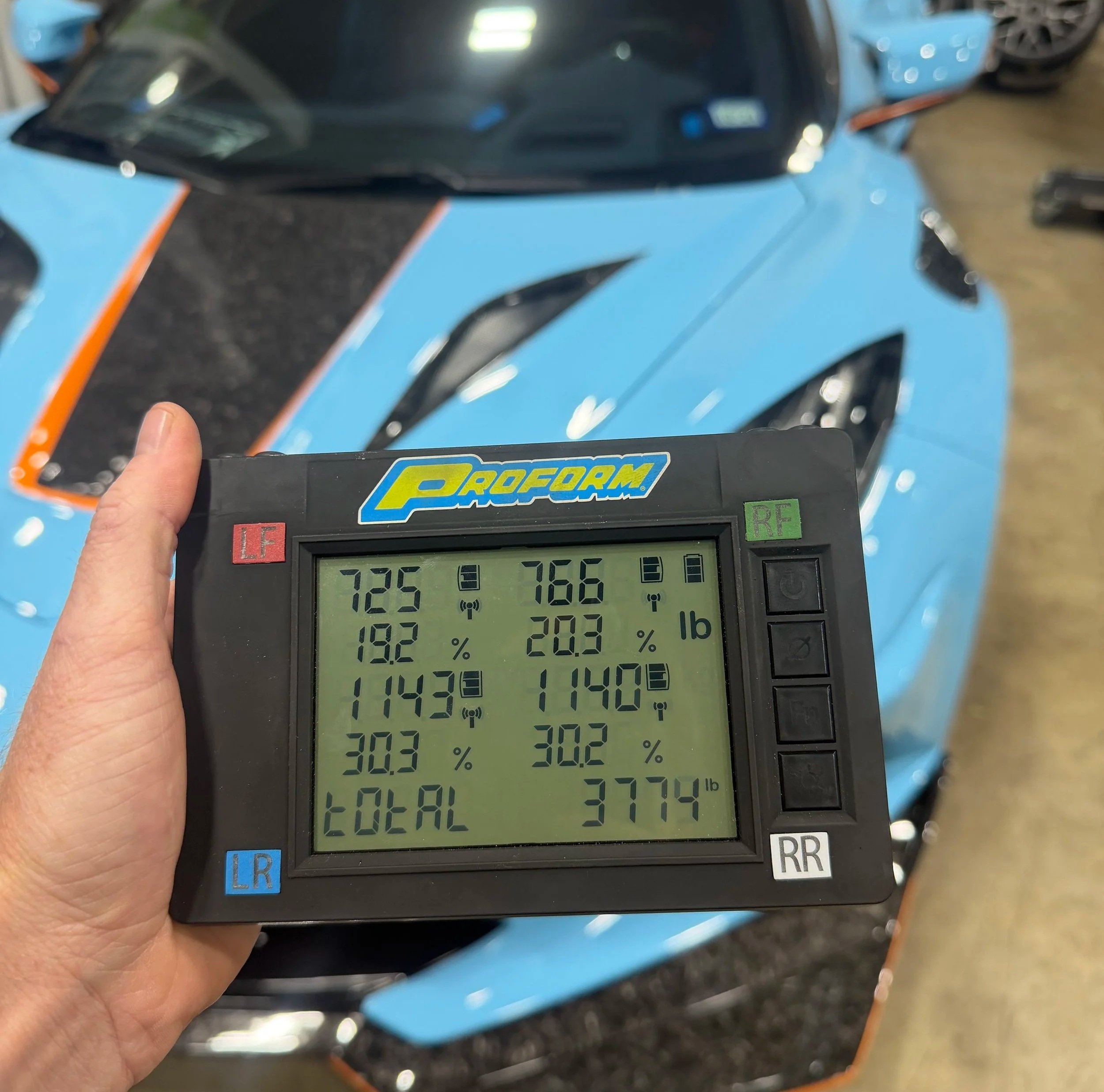 Close-up of a digital weather station display showing temperature, humidity, and other weather data, with a blurred background of a blue motorcycle.