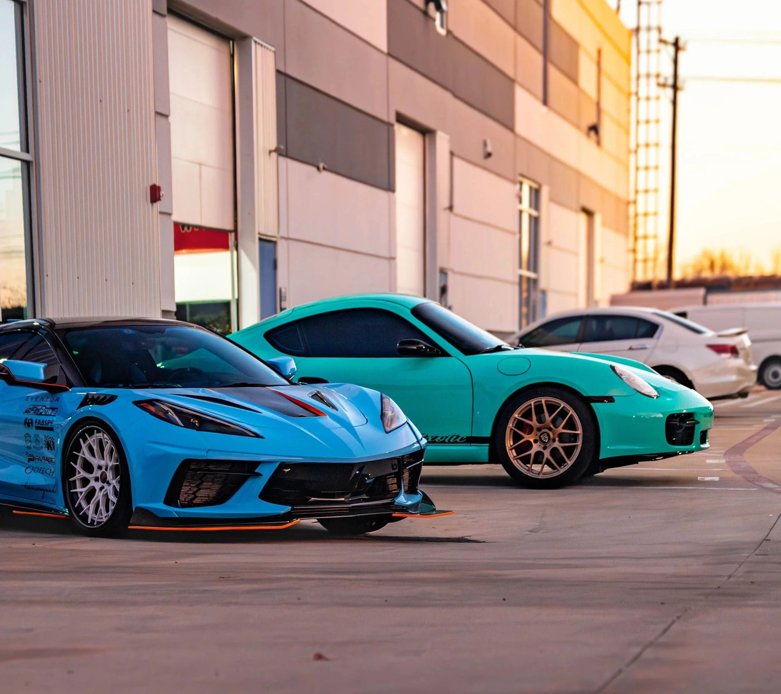 Several sports cars parked in a lot outside a modern building at sunset, with clear skies and warm lighting.