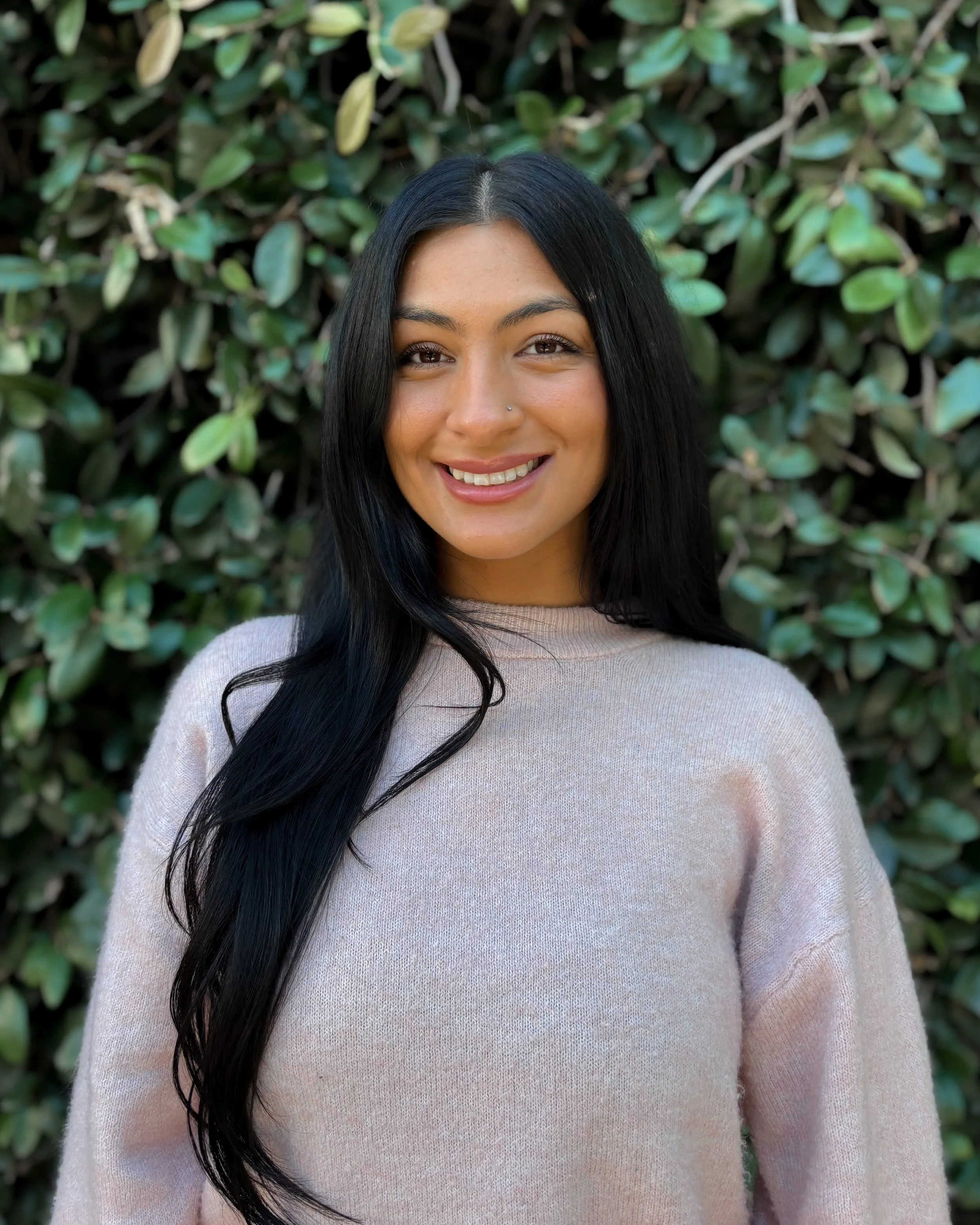 A woman with long black hair smiling in front of green leafy bushes, wearing a beige sweater.