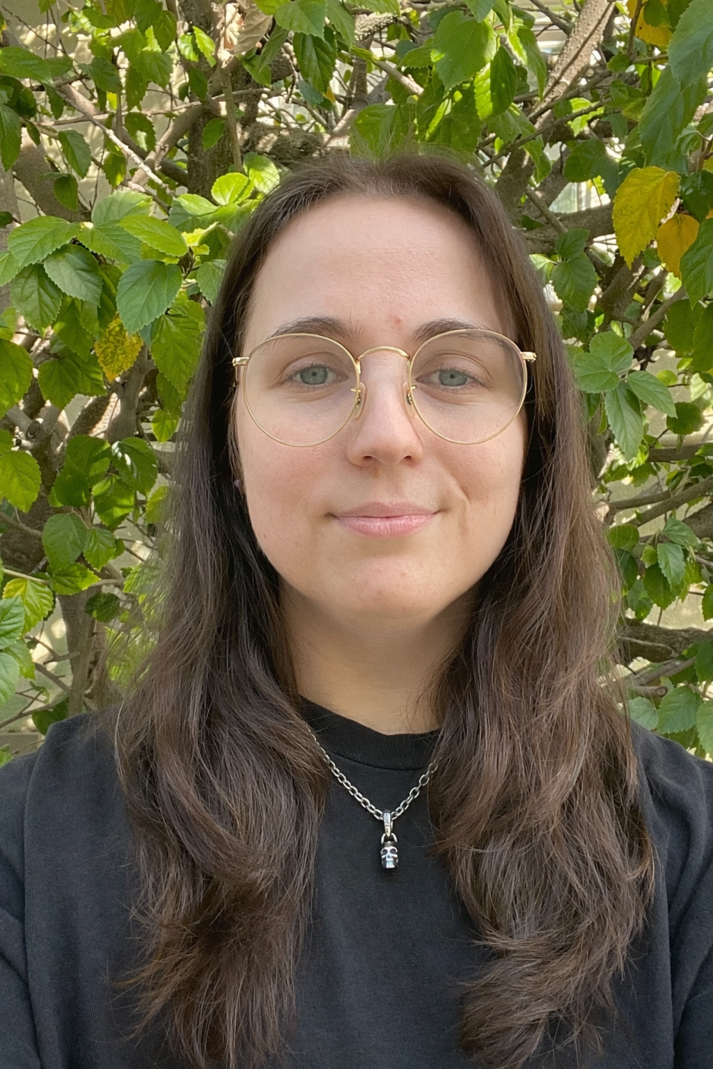 A young woman with long brown hair wearing glasses and a black shirt standing outdoors in front of leafy green shrubbery.
