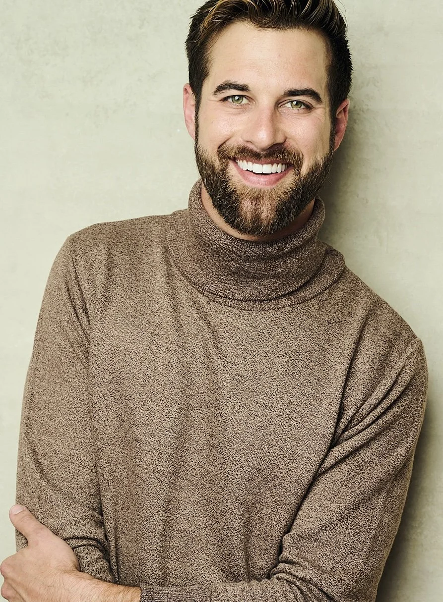 A smiling man with a beard and short dark hair, wearing a brown turtleneck sweater, standing against a light-colored textured wall.
