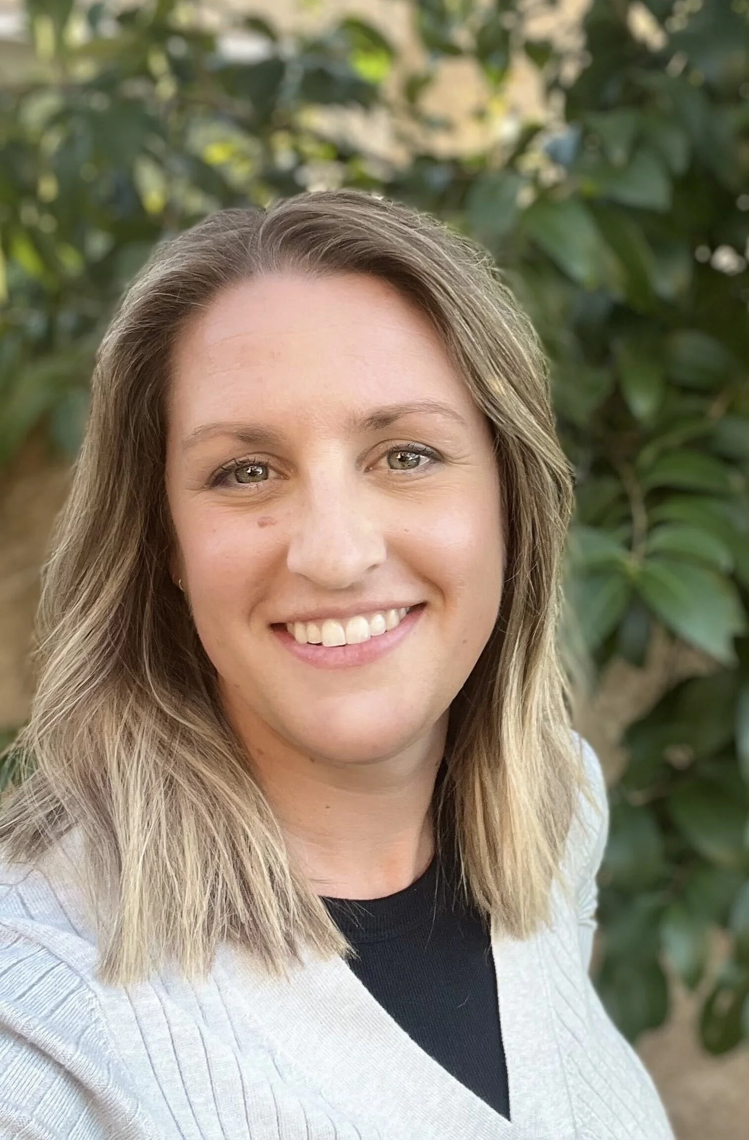A young woman with blonde hair and green eyes smiling outdoors with green foliage in the background.