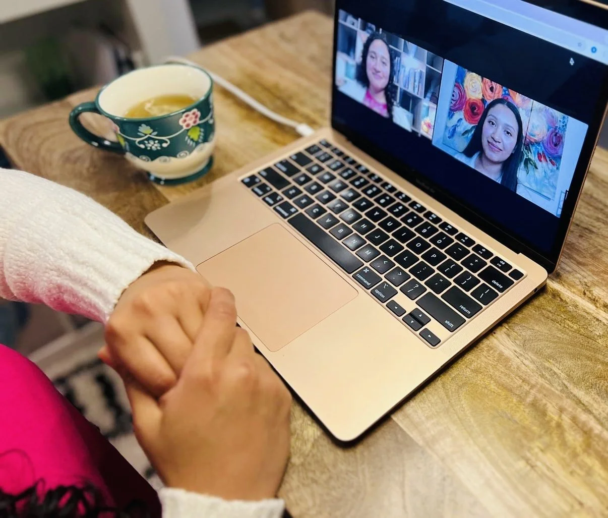 Person holding a child's hand while using a laptop for a video call, with a floral-patterned mug of tea on the wooden table.