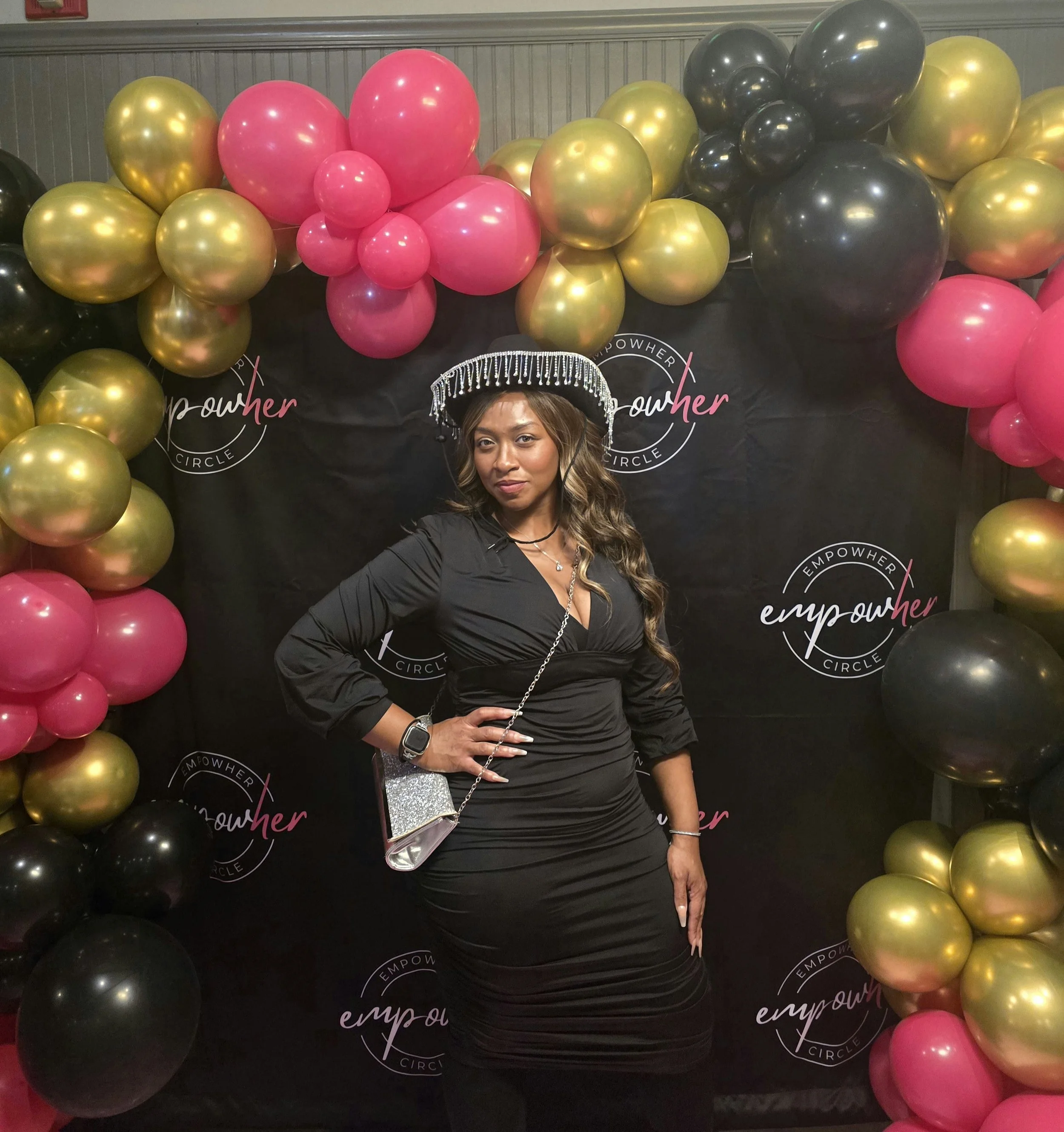 A woman in a black dress poses in front of a backdrop surrounded by pink, black, and gold balloons at an empowerment event.