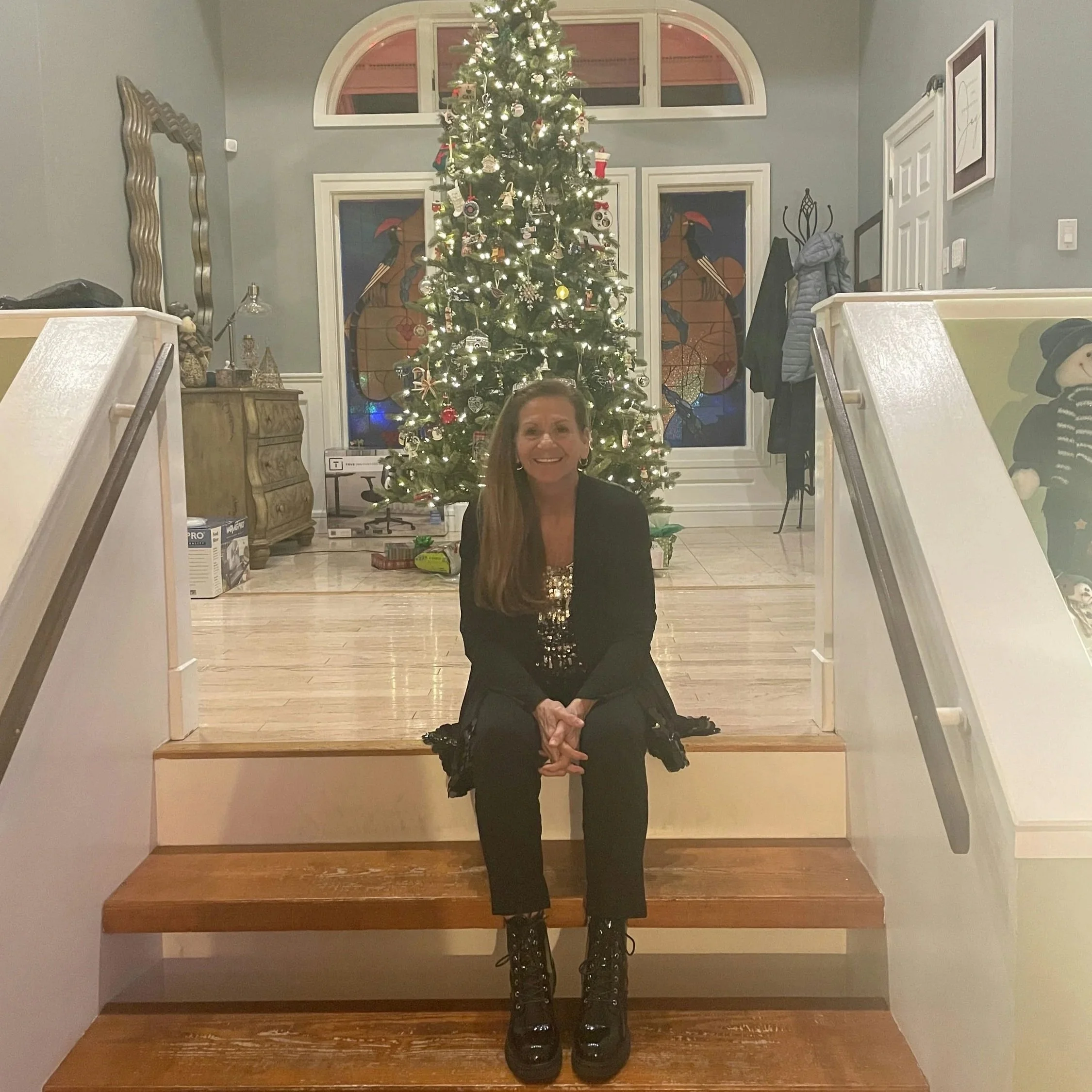 Woman sitting on stairs in front of a decorated Christmas tree inside a home.