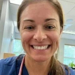 Close-up of a woman smiling, indoors with large windows behind her.