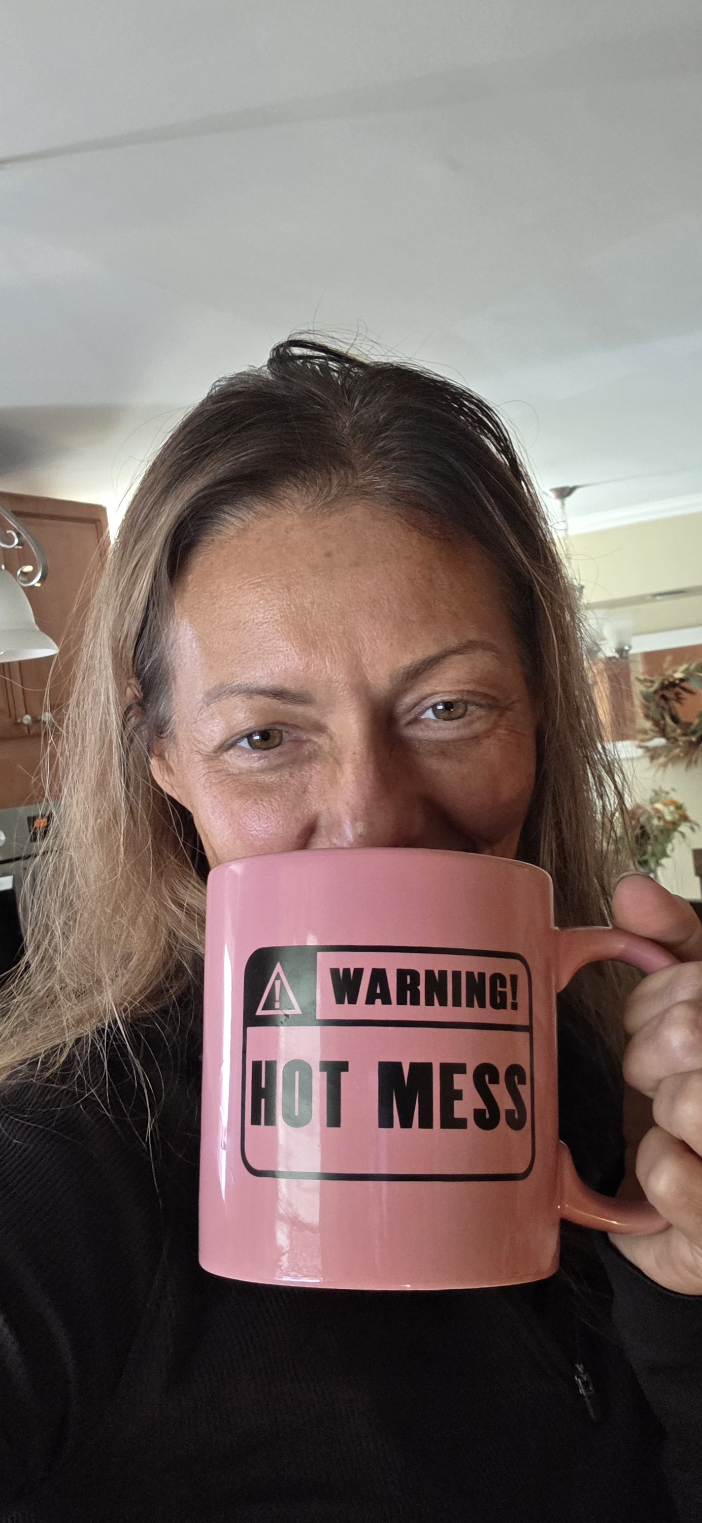 Woman holding a pink mug with a warning label that reads 'WARNING! HOT MESS' in a kitchen setting.