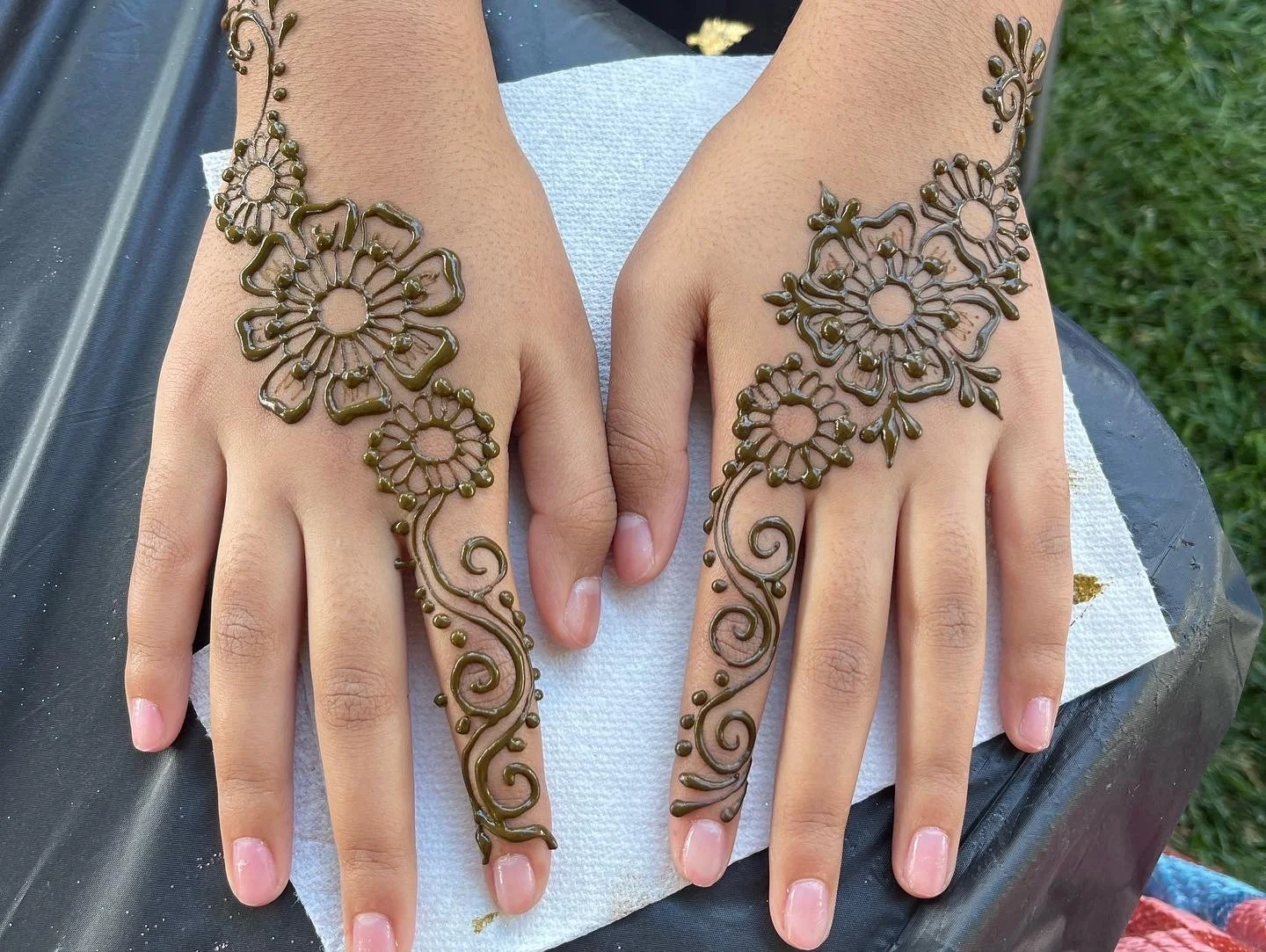 Two hands with henna designs featuring flowers and swirling patterns on fingers, arms, and back of hands.