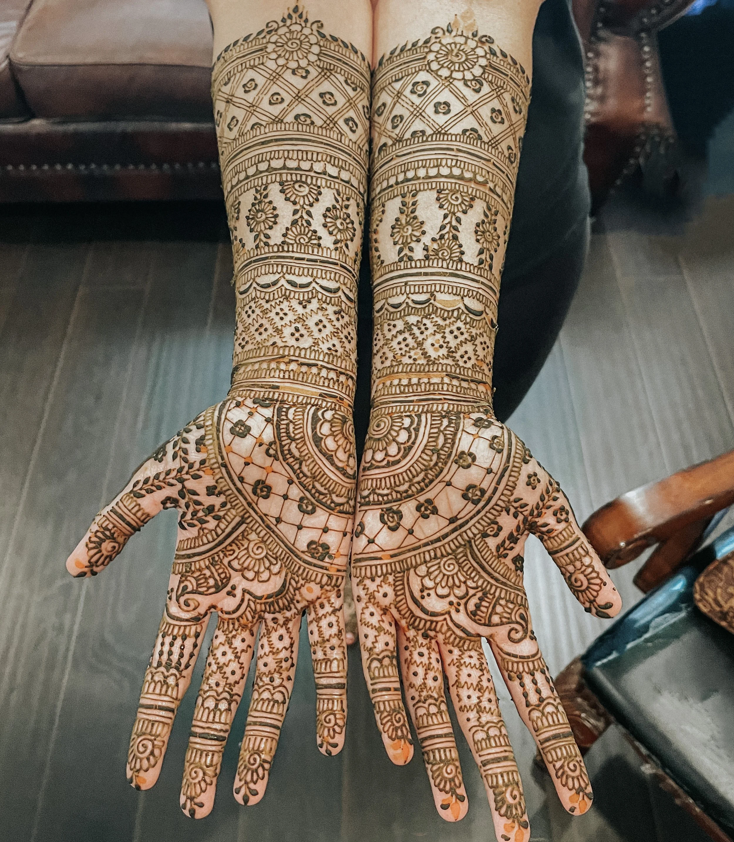 Intricate brown mehndi (henna) designs on the palms and extending up the forearms of two hands, with floral and geometric patterns.