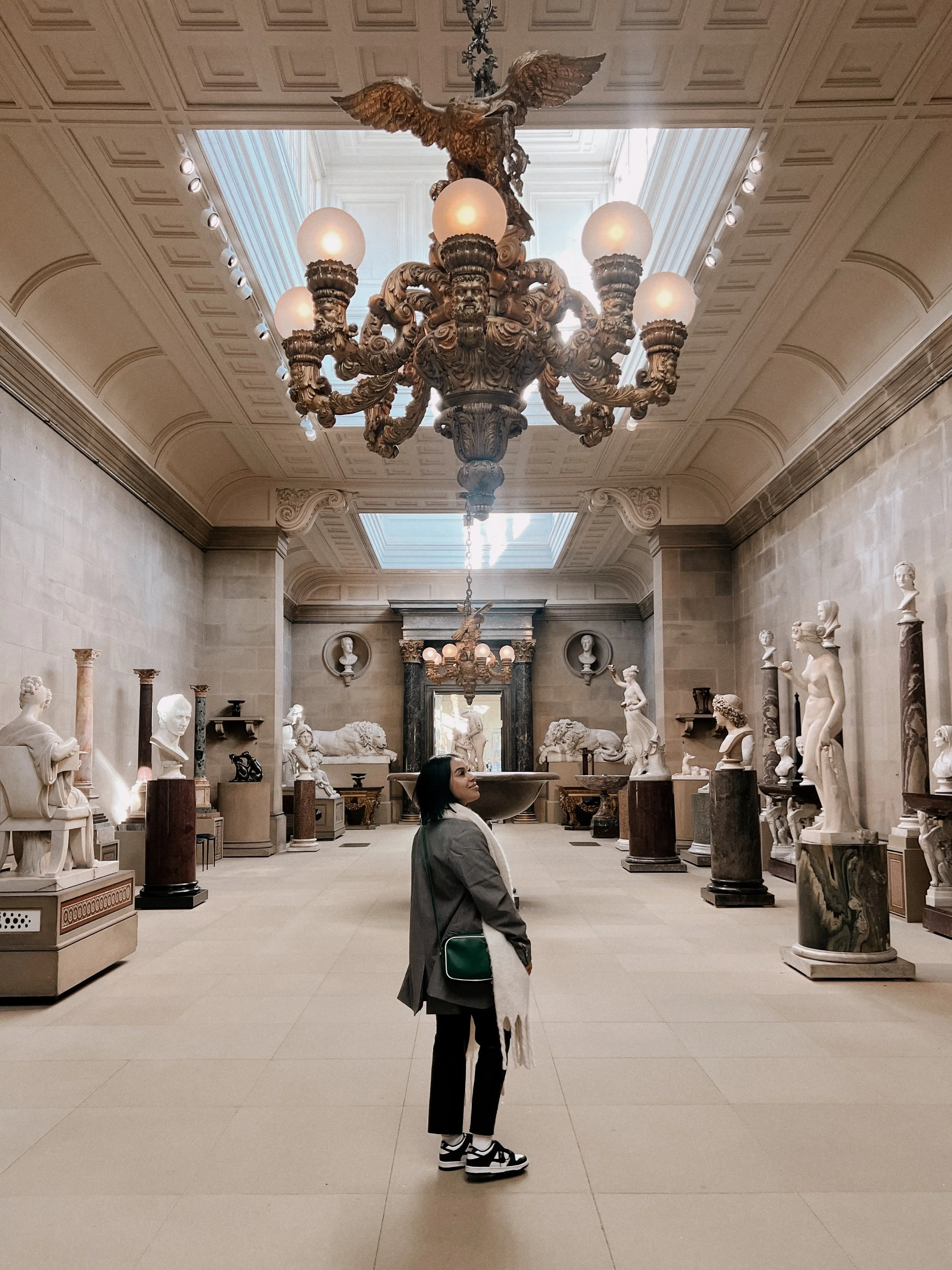 A woman standing in an art gallery or museum with classical sculptures and busts on pedestals, ornate ceiling, and a large chandelier overhead.