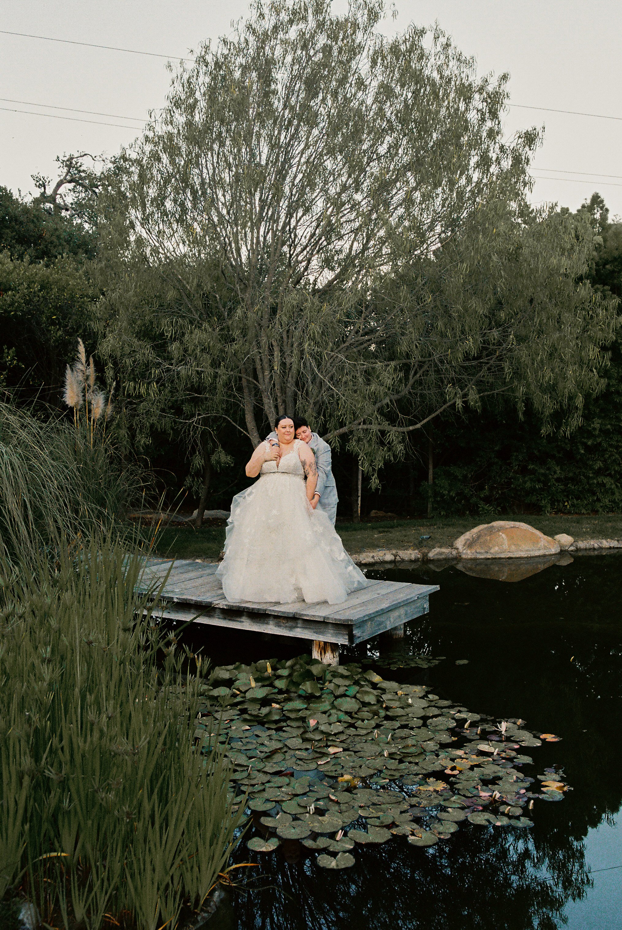 Two women, one in a wedding dress and the other in a suit, standing close together on a wooden dock by a pond with water lilies, surrounded by trees and greenery.
