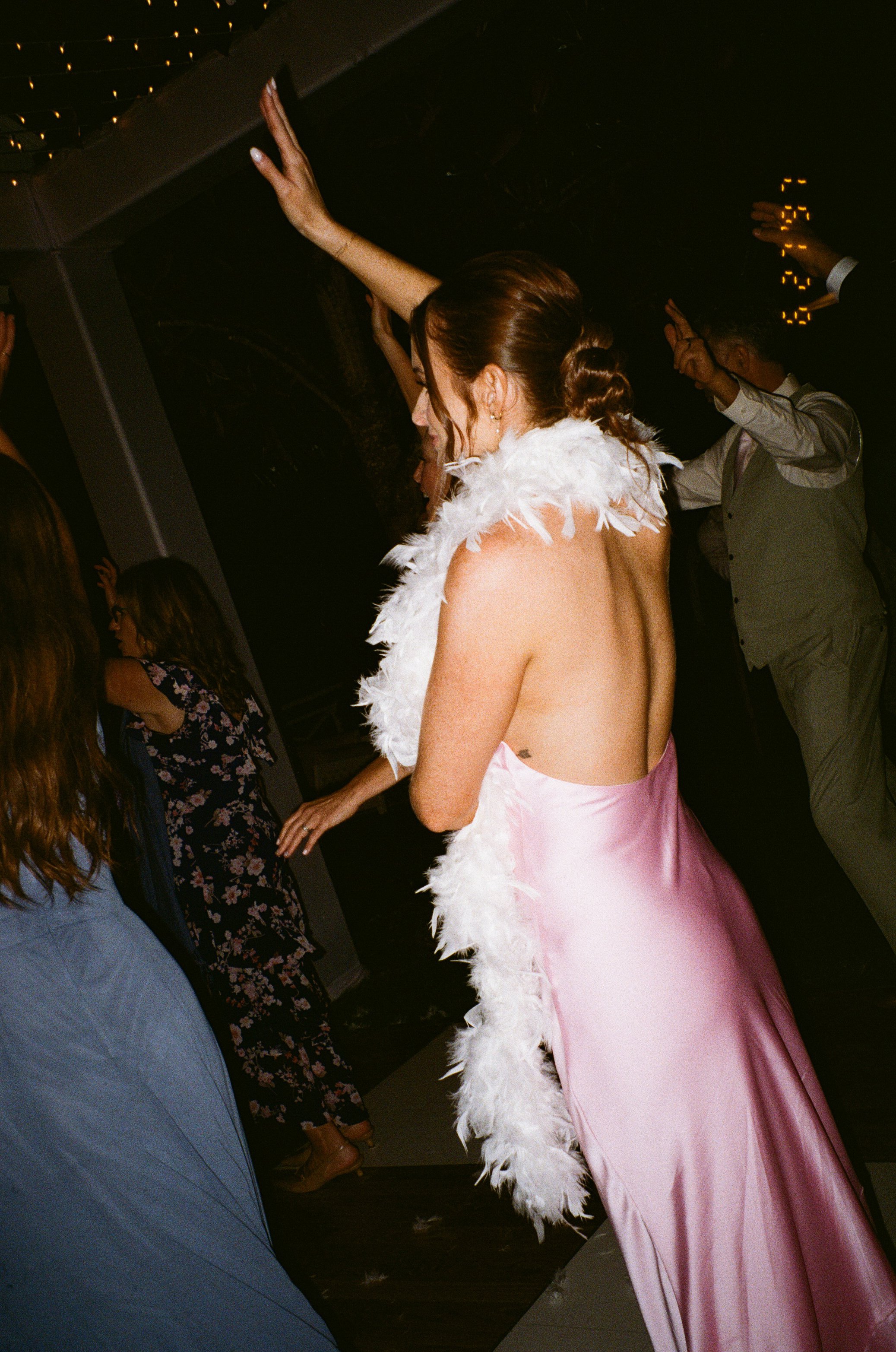 Woman in a pink satin dress with a white feather boa dancing at a party or celebration in a dimly lit room.